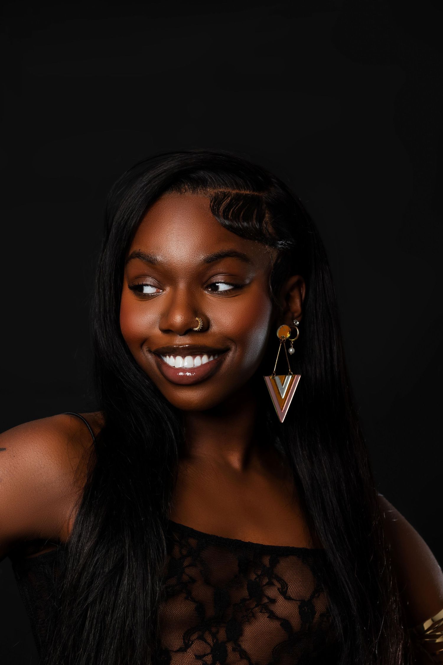 A smiling Black woman with sleek hair and geometric earrings poses against a dark black background.