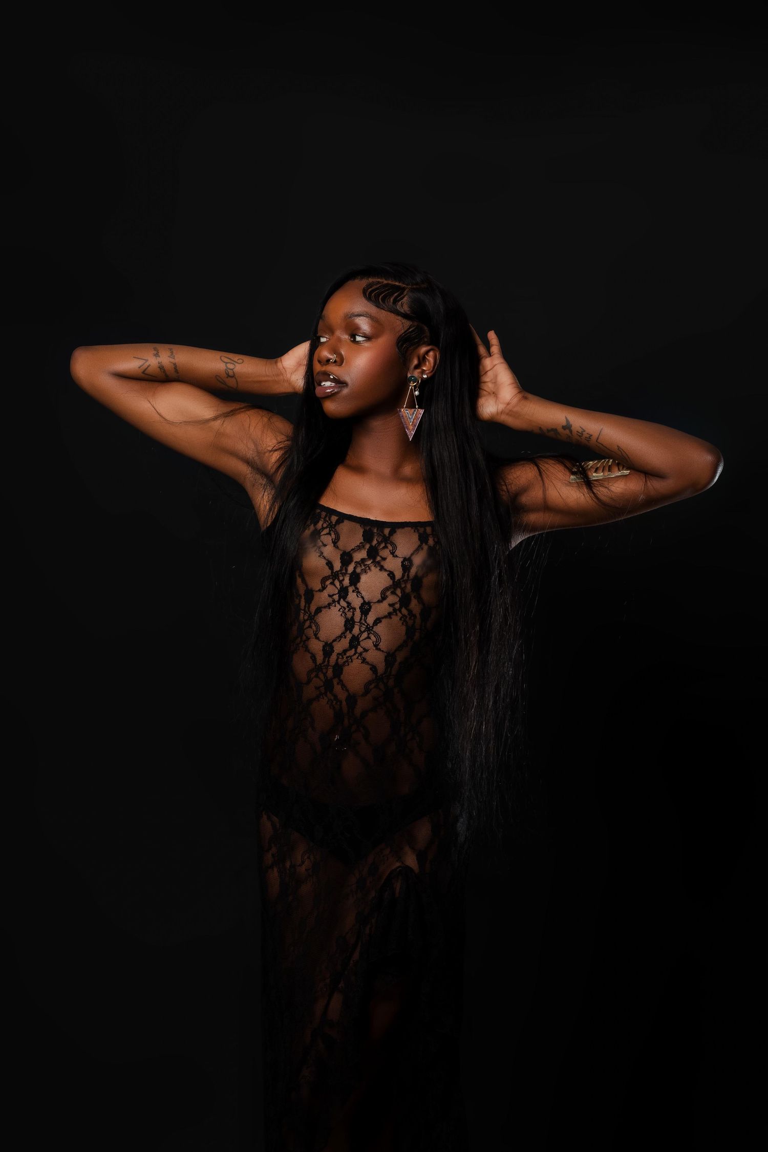 Woman in black lace dress with long straight hair and tattoos posing against dark background.
