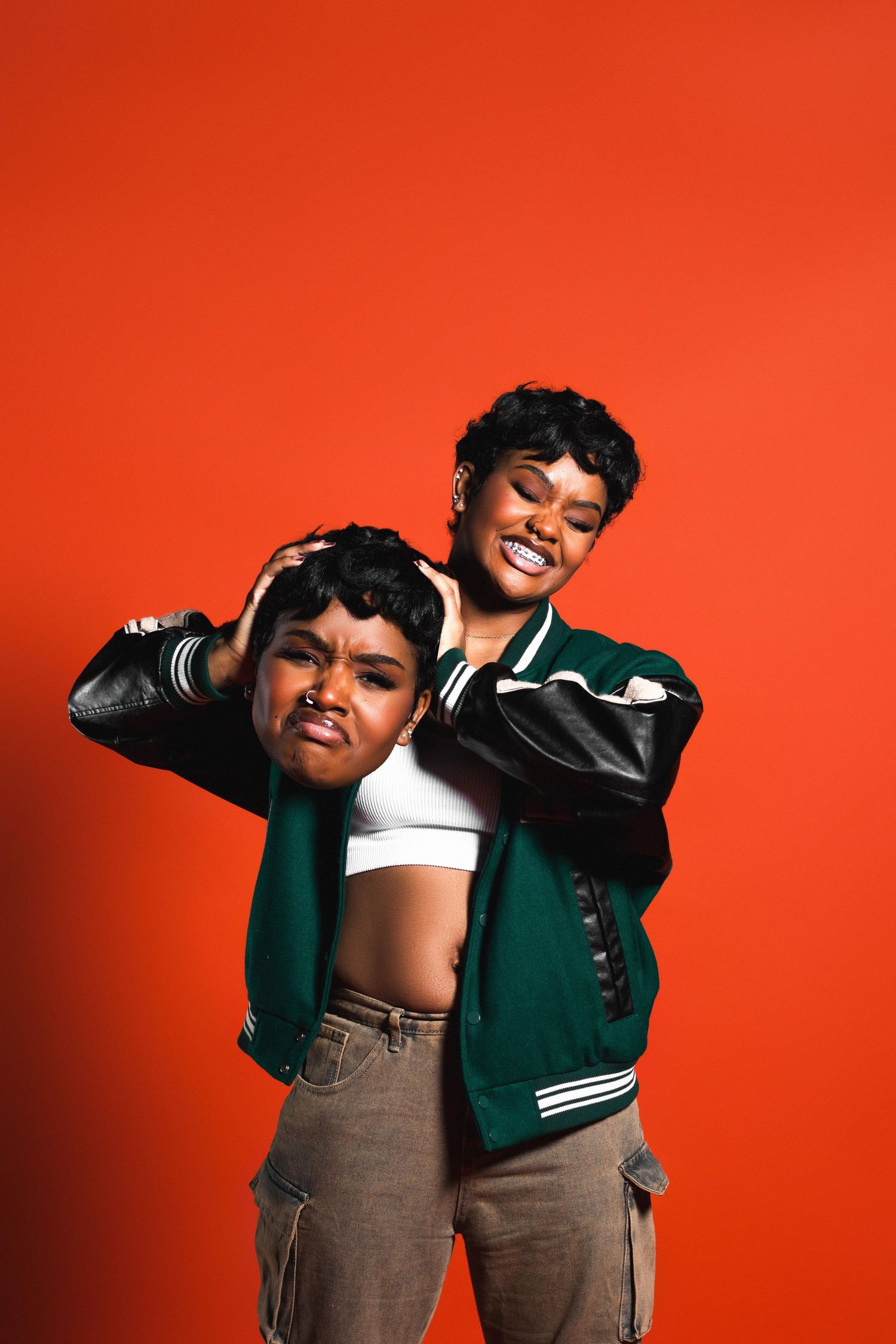 Two young women in green varsity jackets posing playfully against a bright orange-red background.