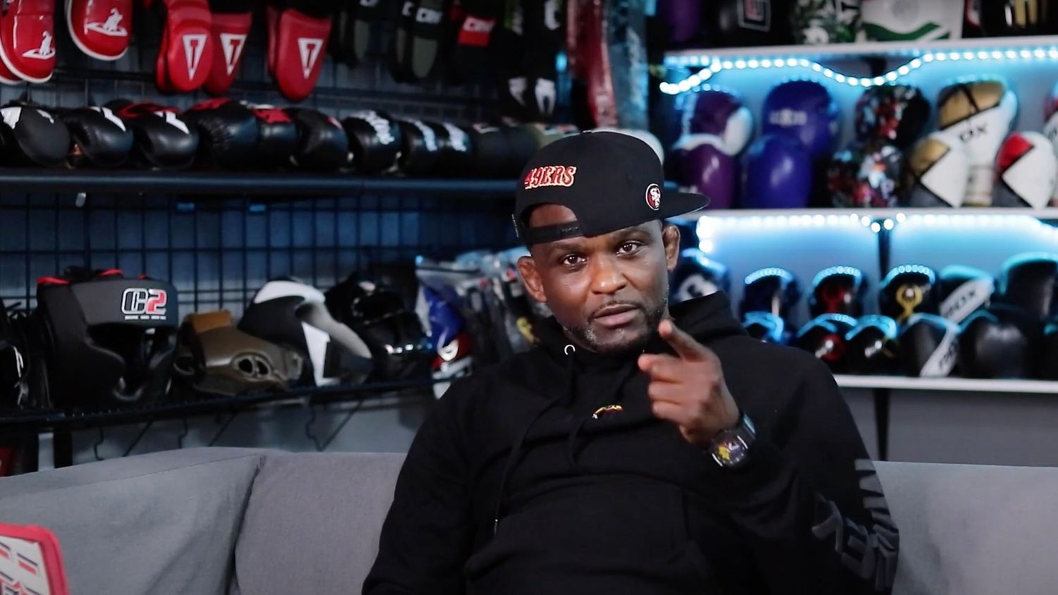Man in black cap sitting in a sports equipment store filled with helmets.