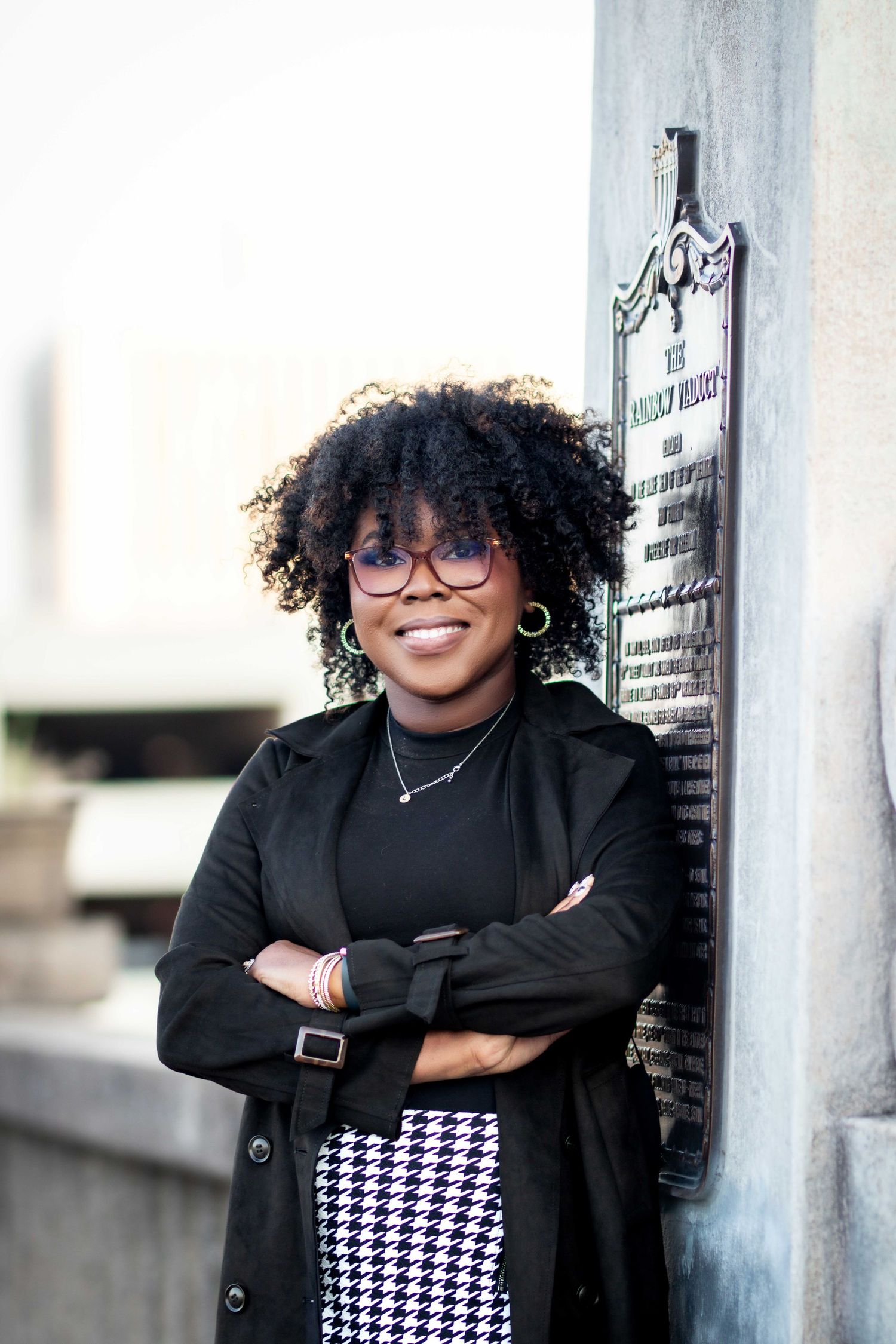 Smiling woman with curly hair and glasses poses confidently against historic plaque.