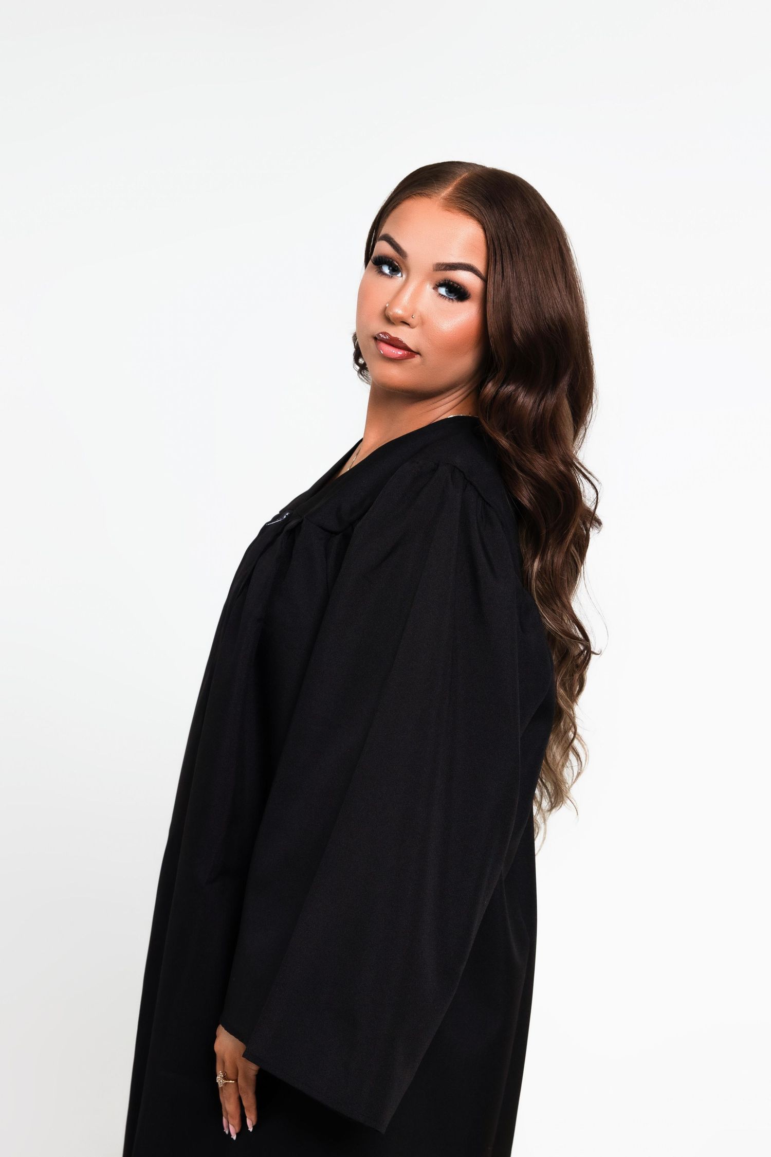 Young woman in black graduation gown with long wavy hair looking over shoulder against white background.