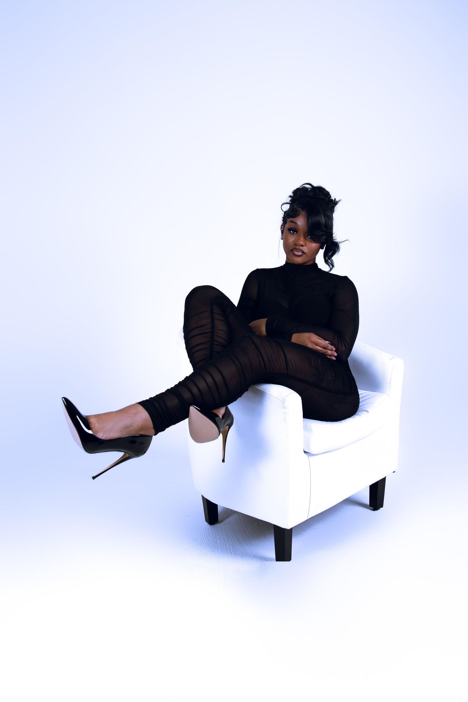 Woman in black sheer outfit seated on white chair with legs crossed wearing stiletto heels.