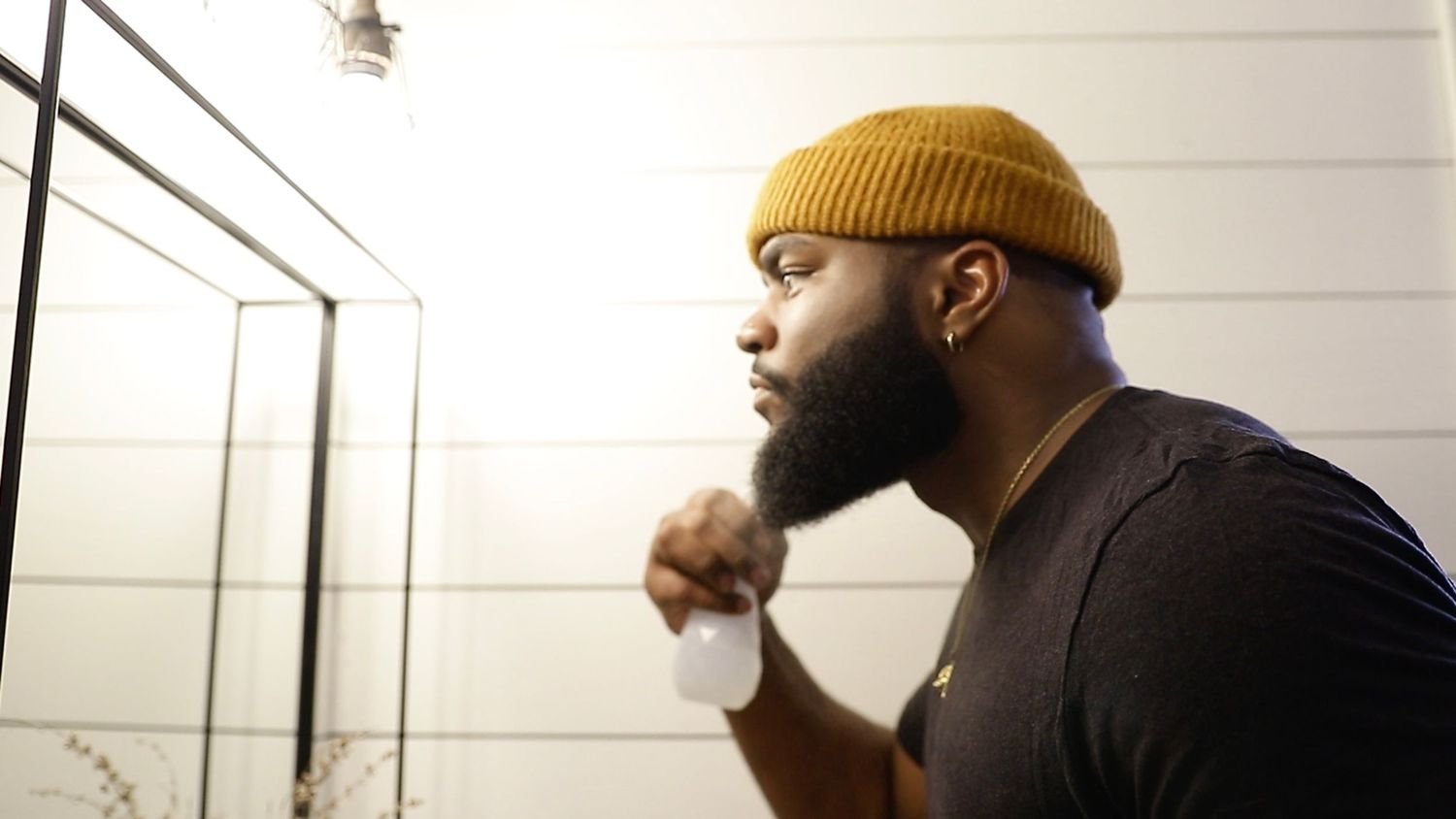 Close-up of bearded man in yellow beanie examining his beard in a bathroom.