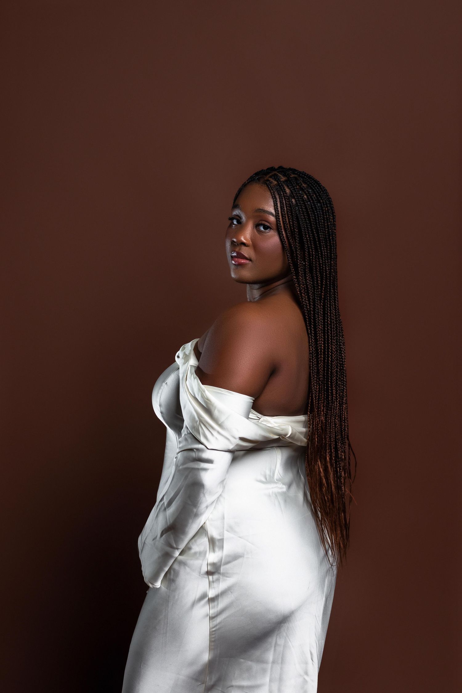 Black woman in white off-shoulder satin dress with long braids, posing sideways against brown background.