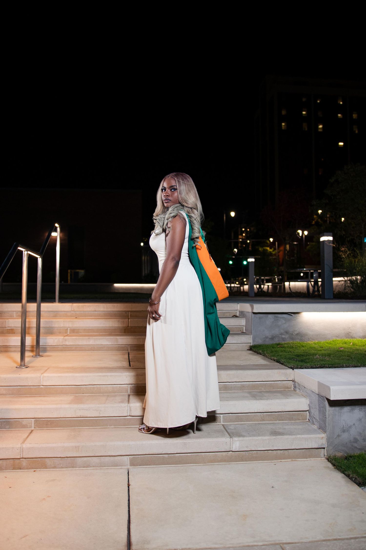 Woman in white maxi dress holding green and orange bag on outdoor steps at night.