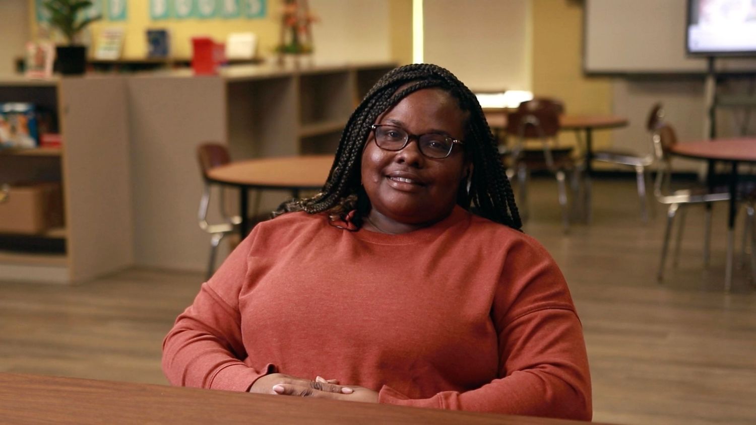Woman in orange sweater smiling while seated in a bright modern classroom.
