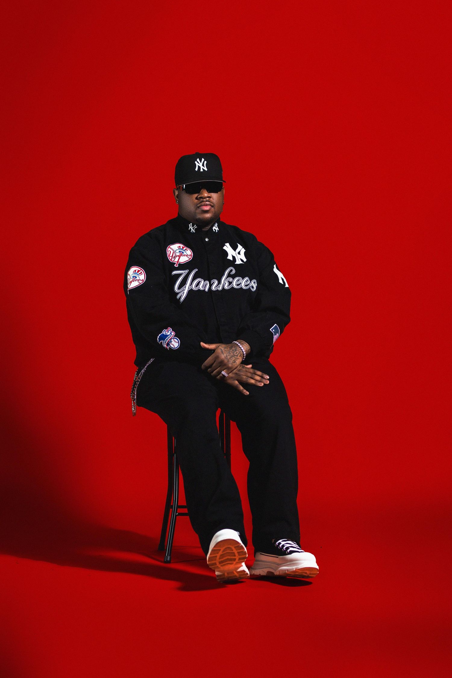 Man in black Yankees jacket and cap sitting on stool against vibrant red background.