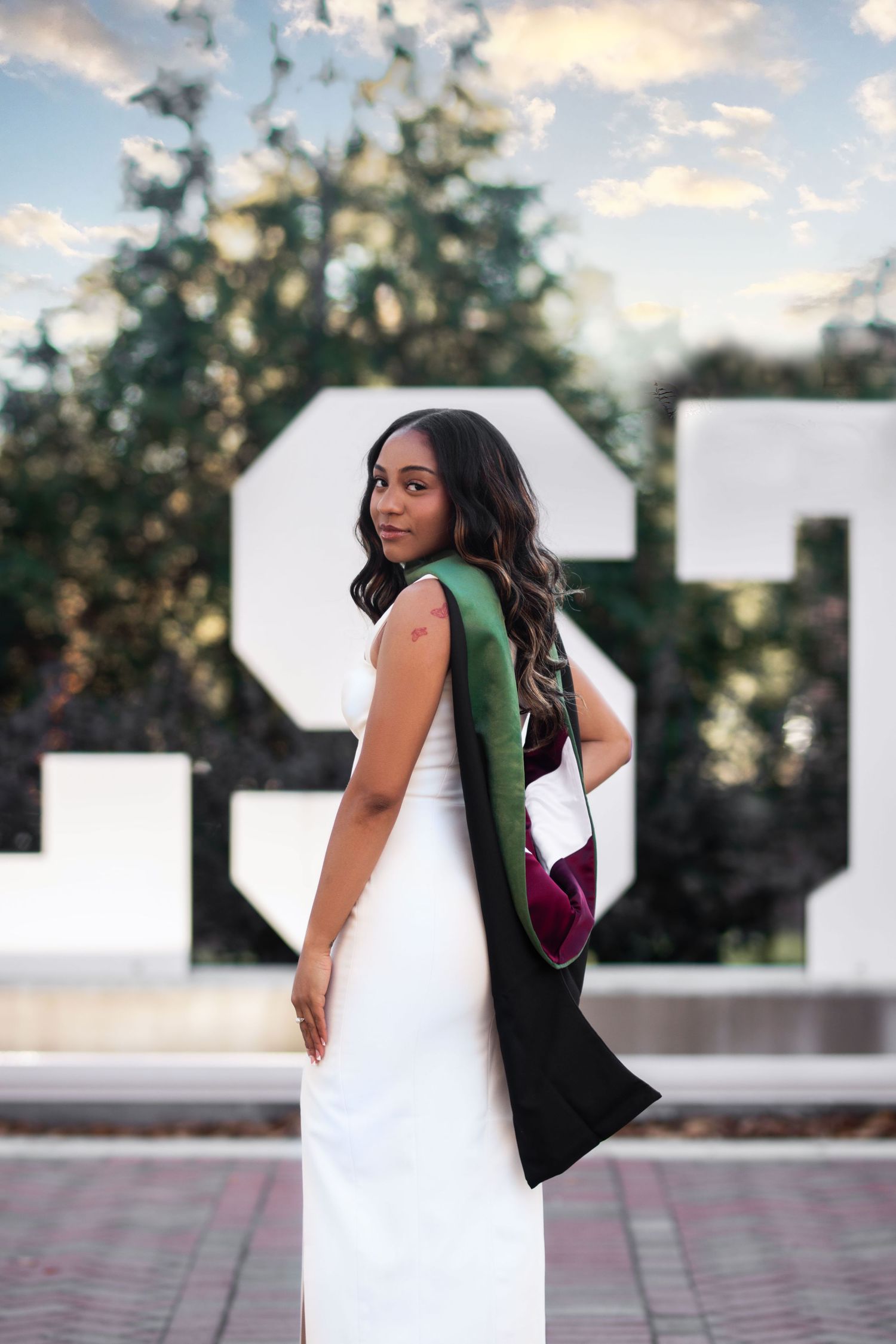 Young Black woman in white dress with green and maroon graduation hood posing near MSU sign.
