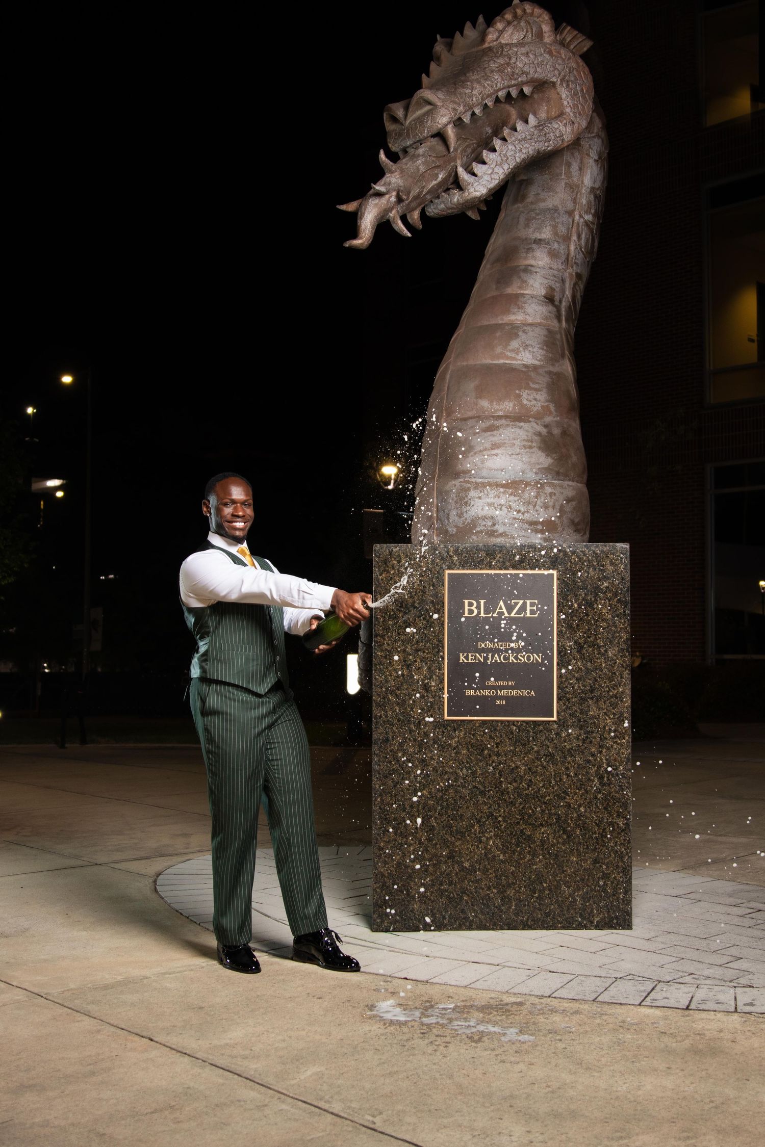 Man in green suit posing with Blaze dragon statue by Ken Jackson at night.