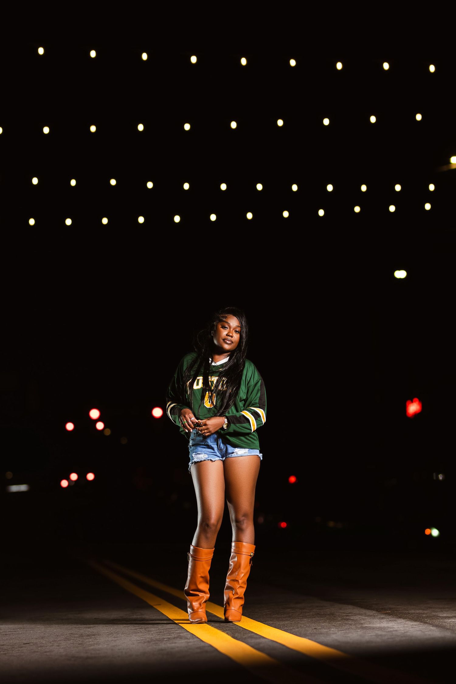 Young Black woman in green varsity jacket, denim shorts, and brown boots standing on a road at night with string lights.