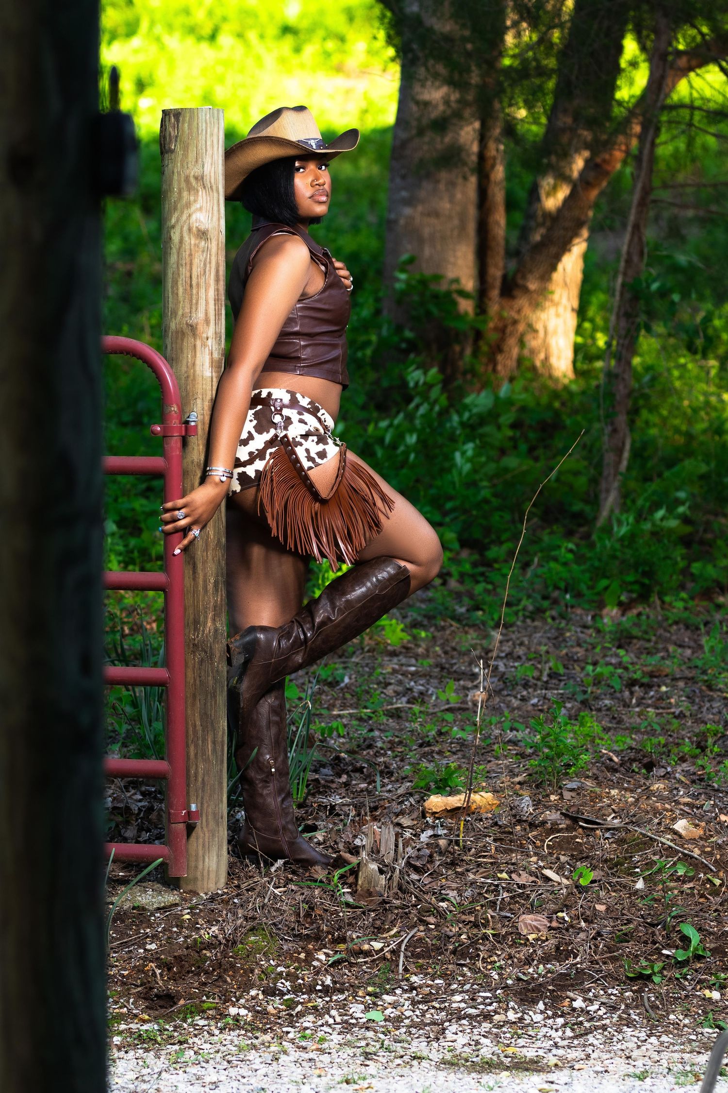 Black woman in cowgirl hat, brown fringe skirt, and boots leaning on a red gate in a wooded outdoor setting.