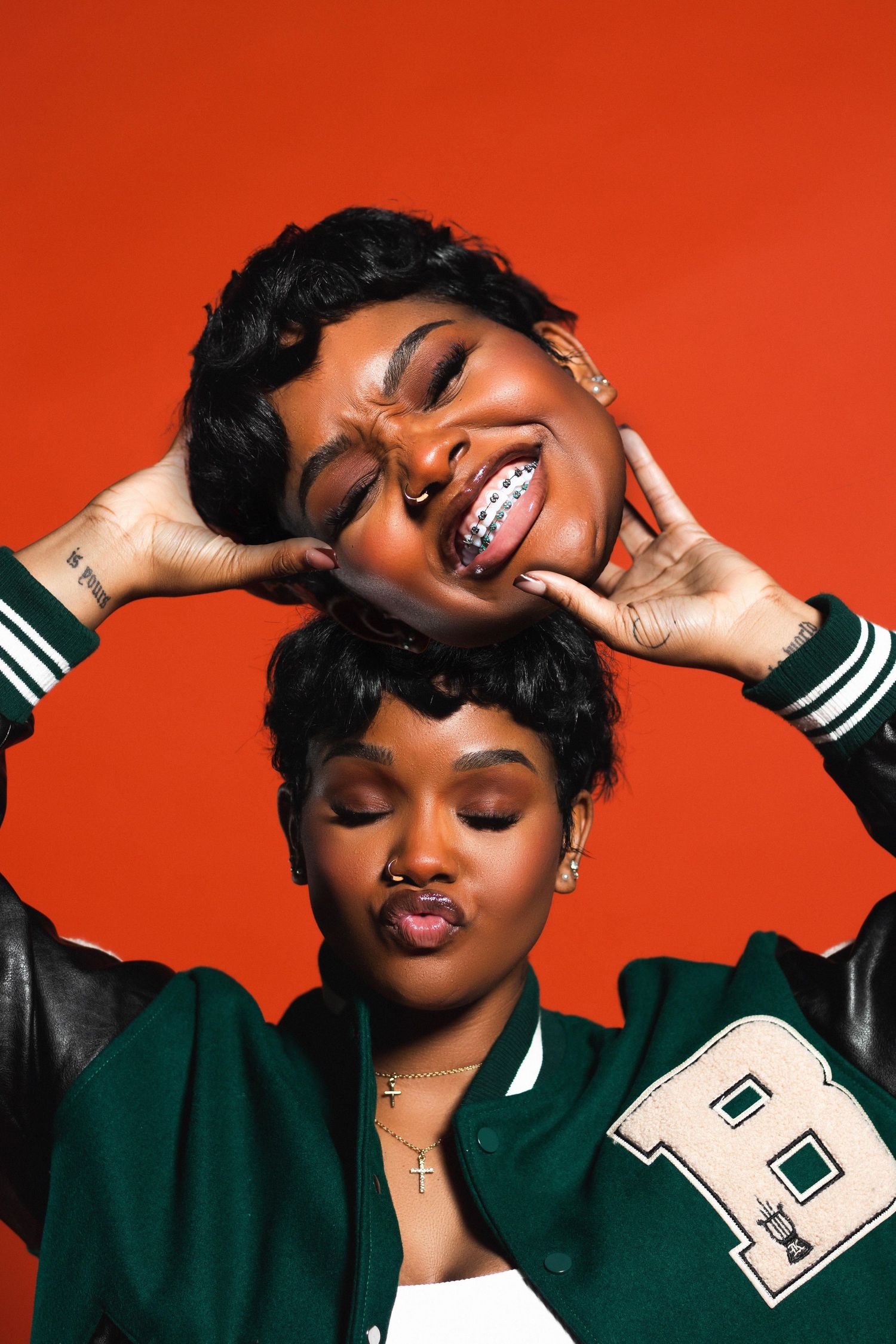 Two young women pose playfully against a bold red background, wearing a green varsity jacket with braces and short hair.