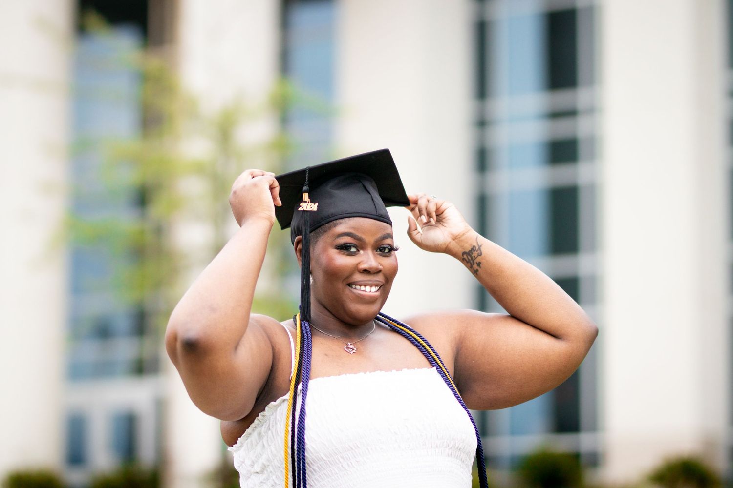 Smiling woman adjusting black graduation cap wearing white dress and colorful kente stole.