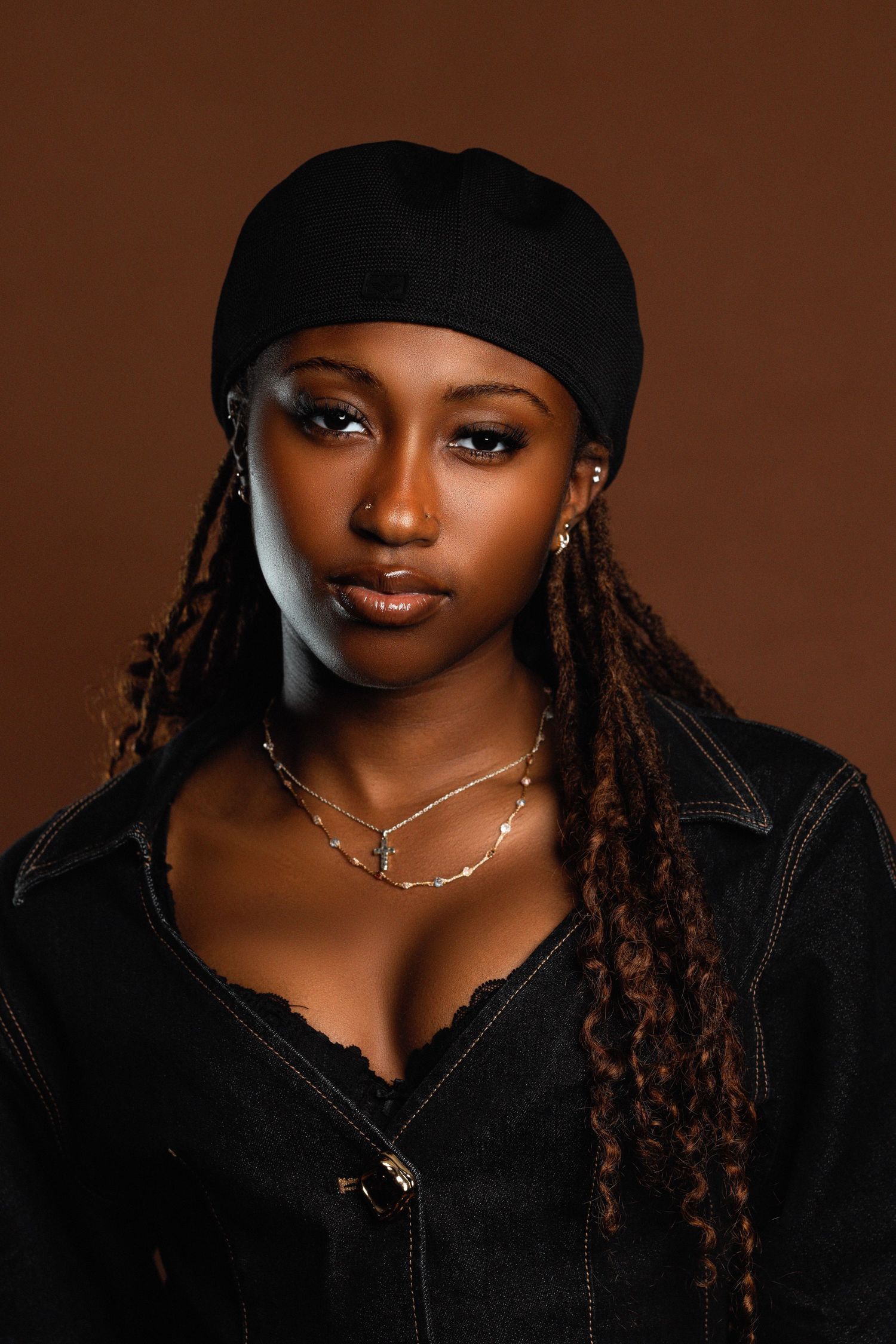 A young Black woman with braids and a black headwrap wears layered gold necklaces and a denim jacket.