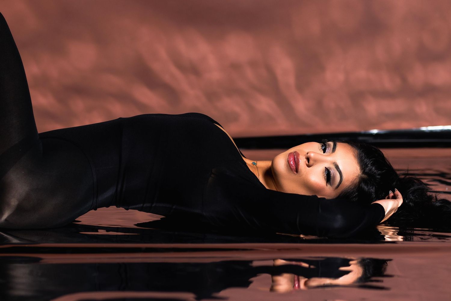 Woman in black bodysuit reclines on reflective floor with warm brown background in studio.
