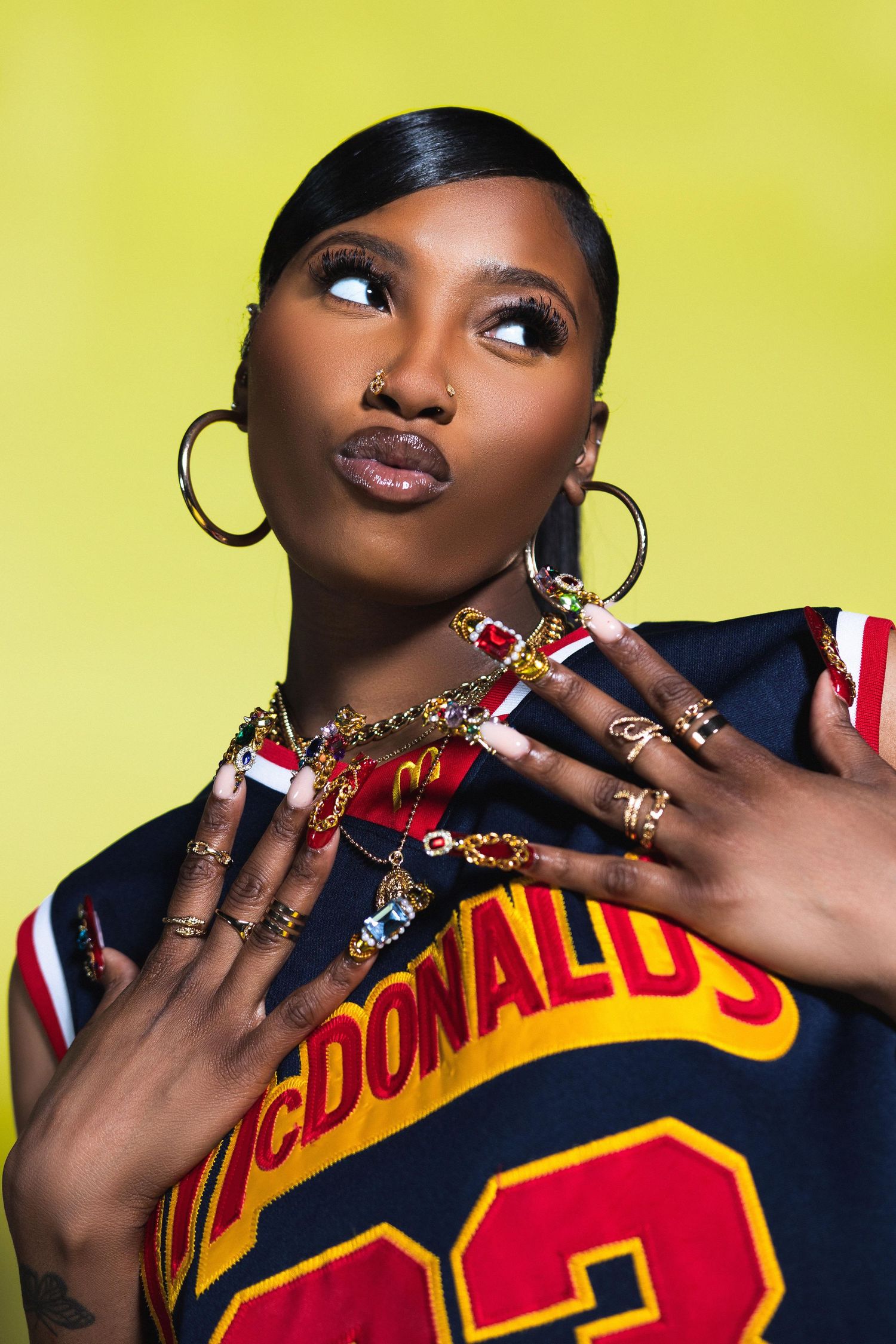 Woman in McDonald's jersey with gold jewelry and long nails poses on yellow background.