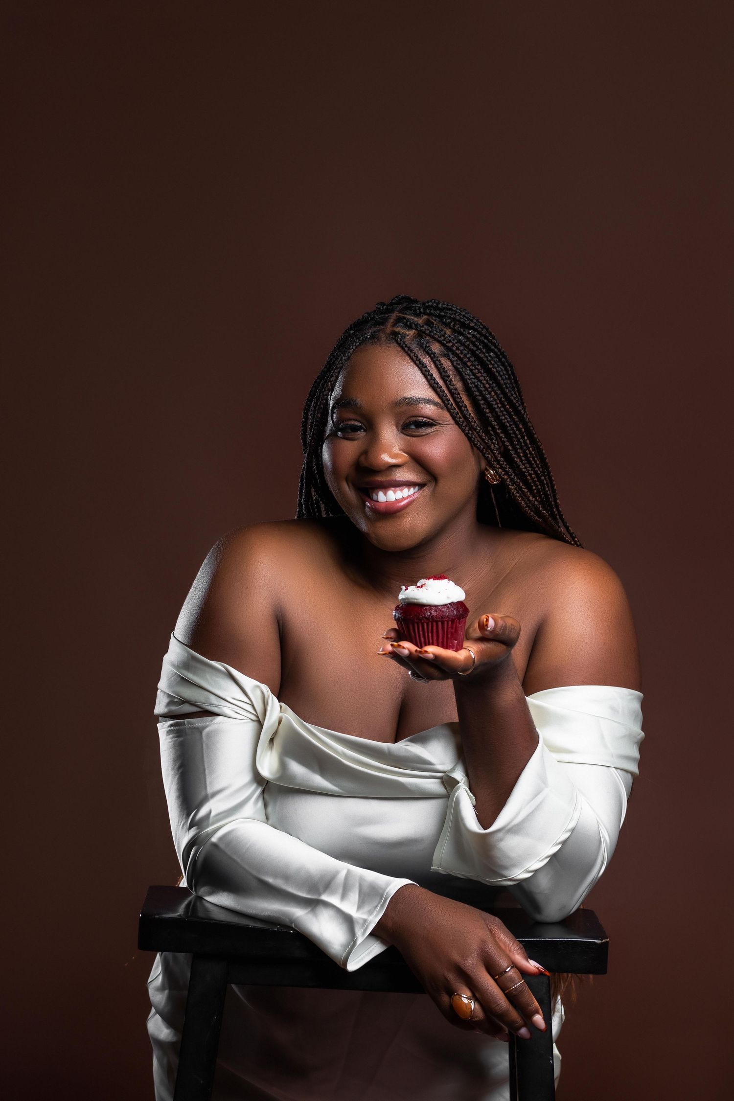 Smiling plus-size woman in white off-shoulder top holding a red velvet cupcake on brown background.