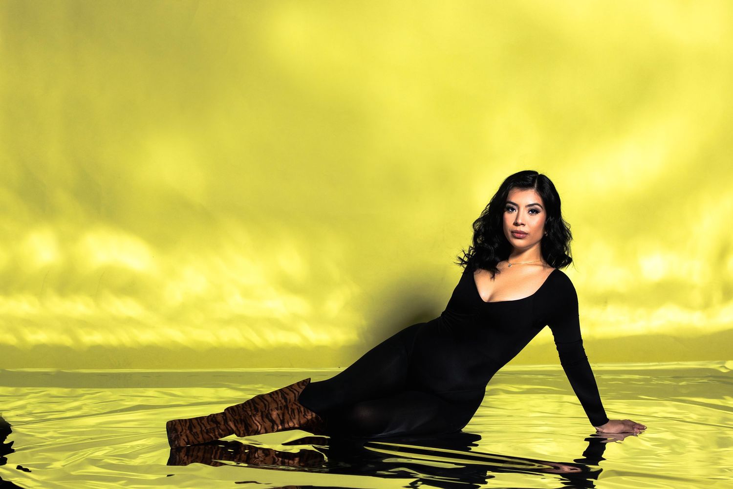 Woman in black dress reclining on reflective surface with yellow backdrop.