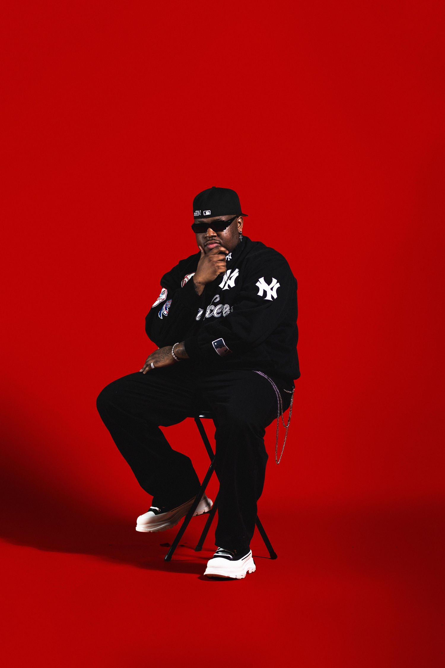 Man in black New York Yankees outfit sitting on chair posing against a bold red background.