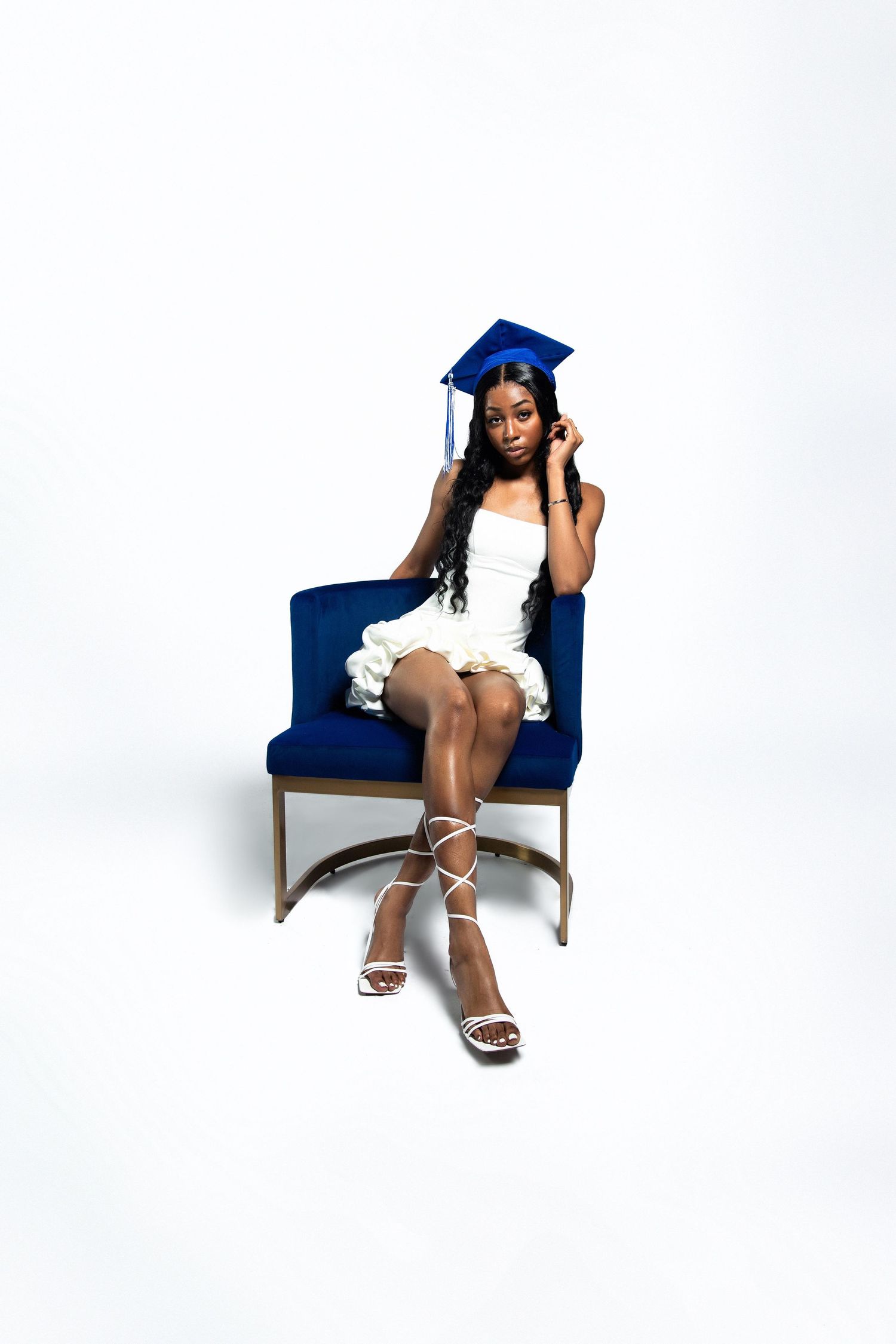Young Black woman in blue graduation cap and white dress seated on a blue velvet chair.