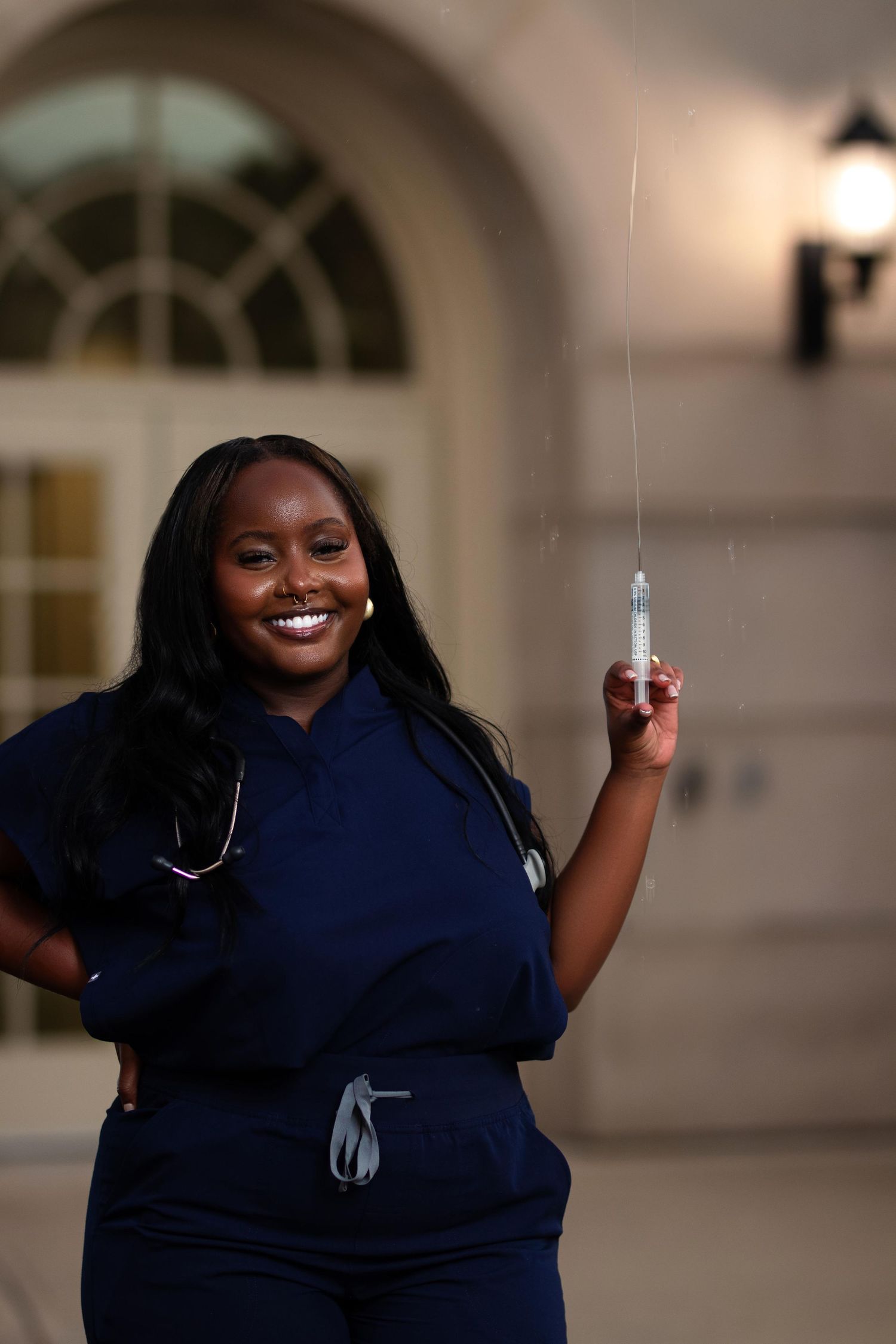 Smiling Black female nurse in navy scrubs holding a syringe outdoors near a building entrance.