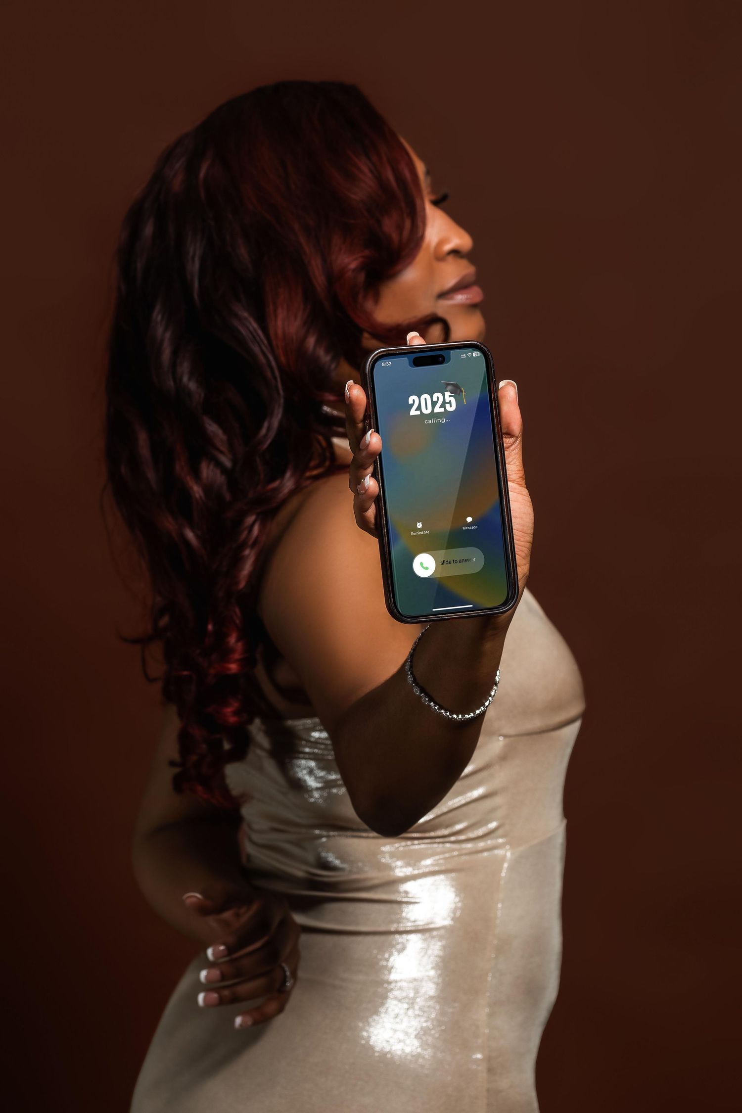 A Black woman with red hair in silver dress holds a phone showing 2025 on a brown background.