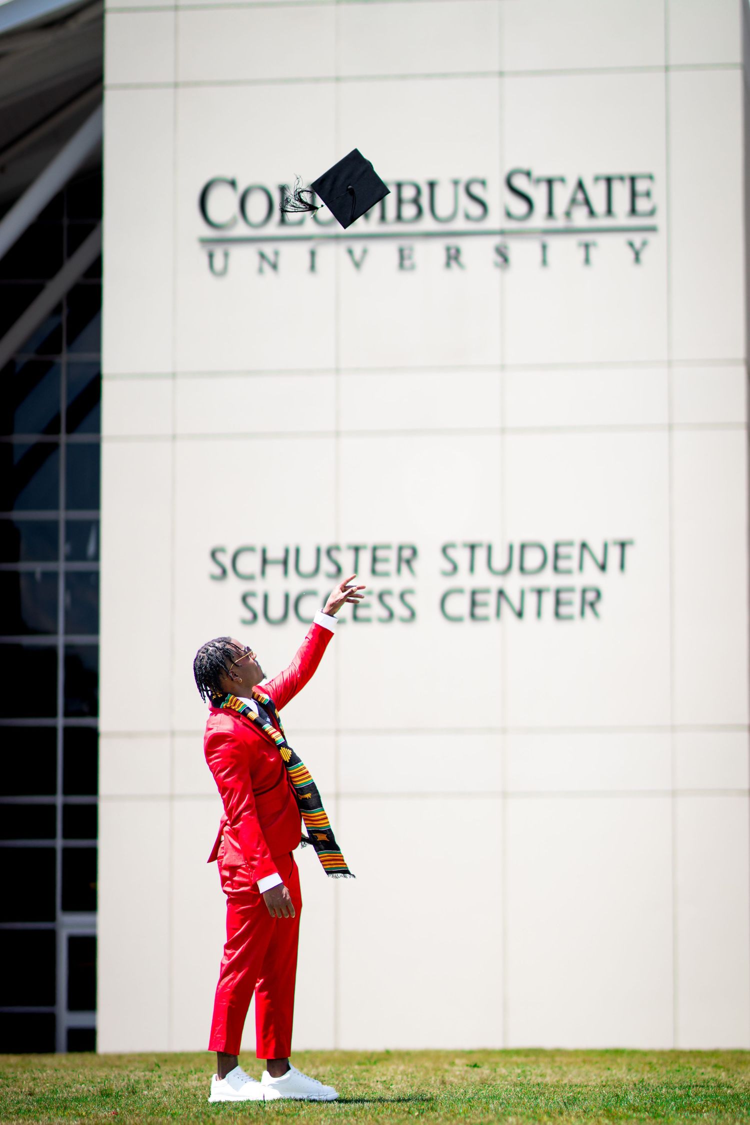 Graduate in red suit tosses cap in front of Columbus State University building.