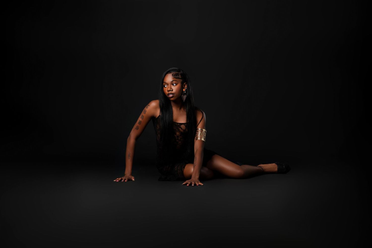 A Black woman in a black outfit poses athletically on a dark studio floor in a dramatic low angle.
