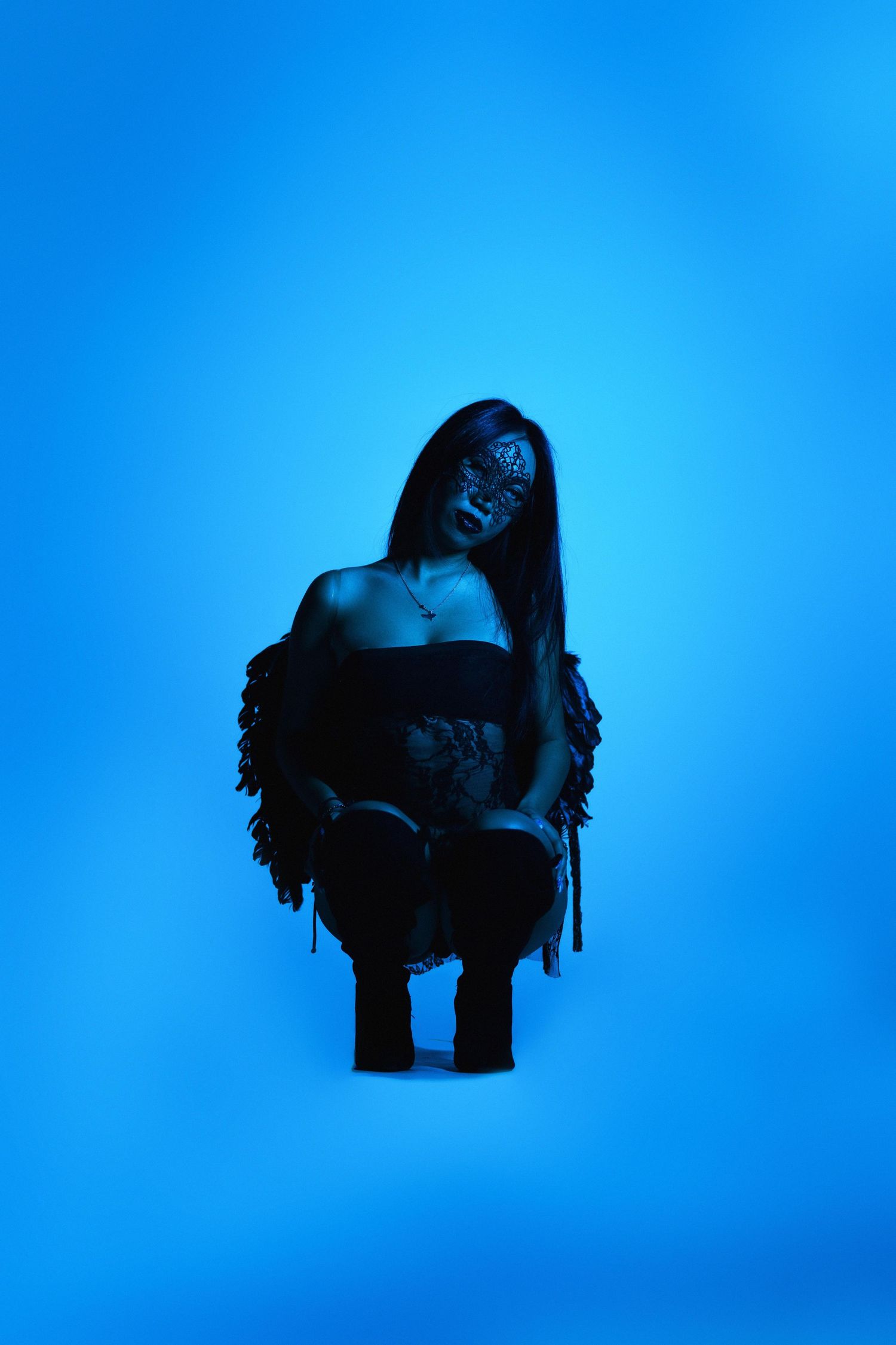 Woman in dark outfit with face paint crouching under blue neon lighting against blue background.