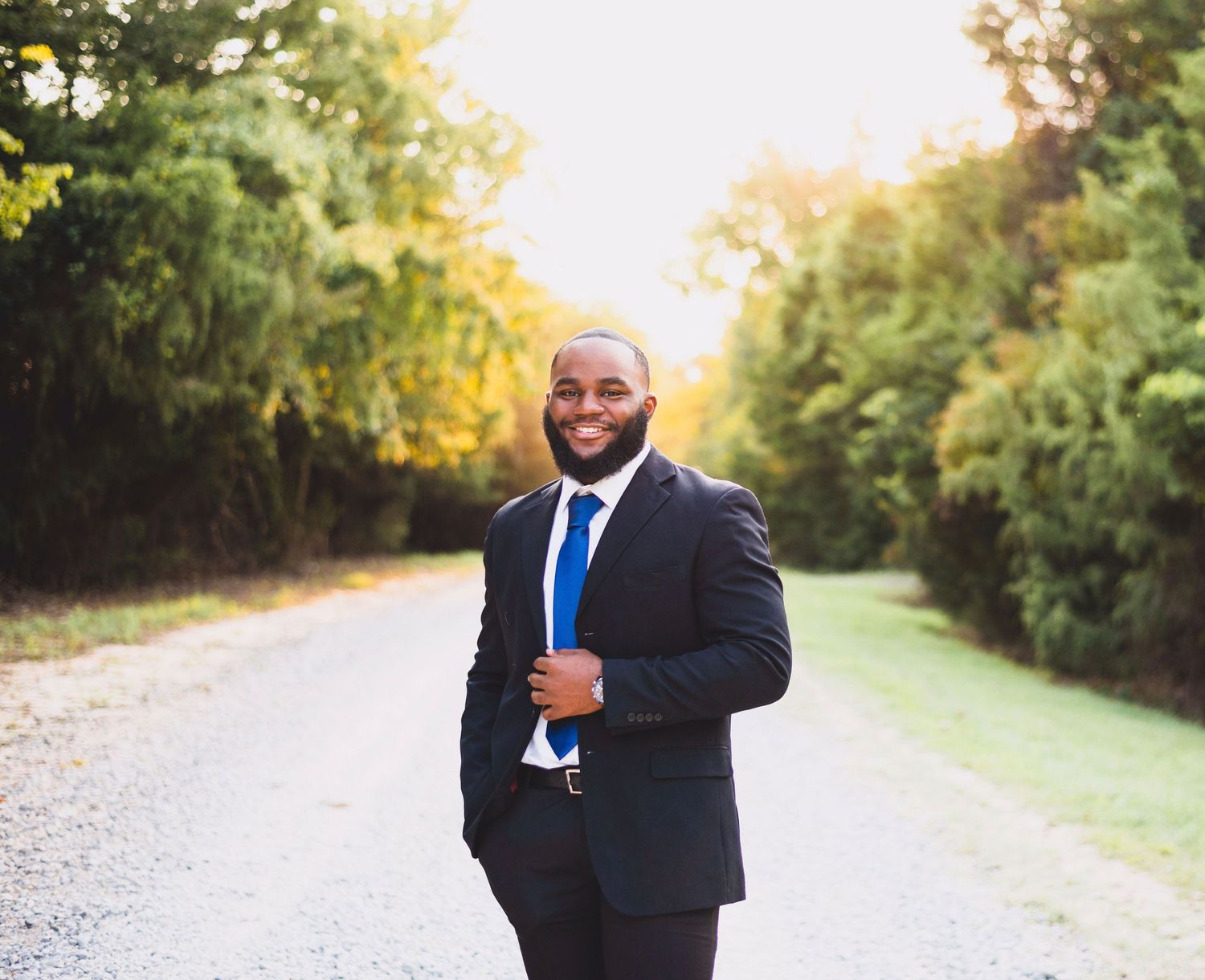 Smiling man in navy suit and blue tie standing on a sunlit country road.