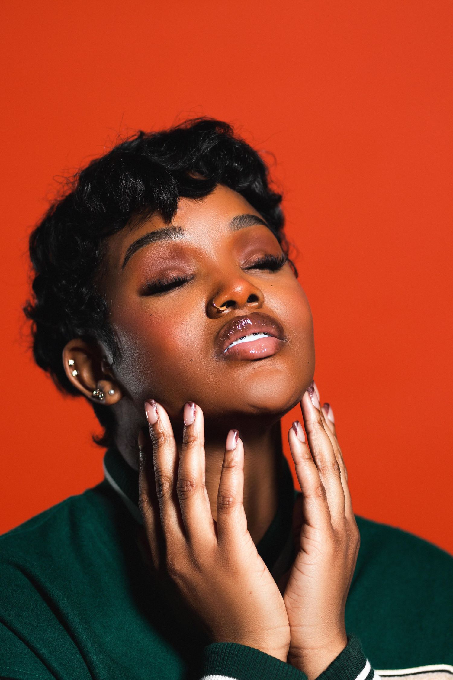 Young Black woman with short curly hair and nose ring, eyes closed, gently touching face with manicured hands on red background.