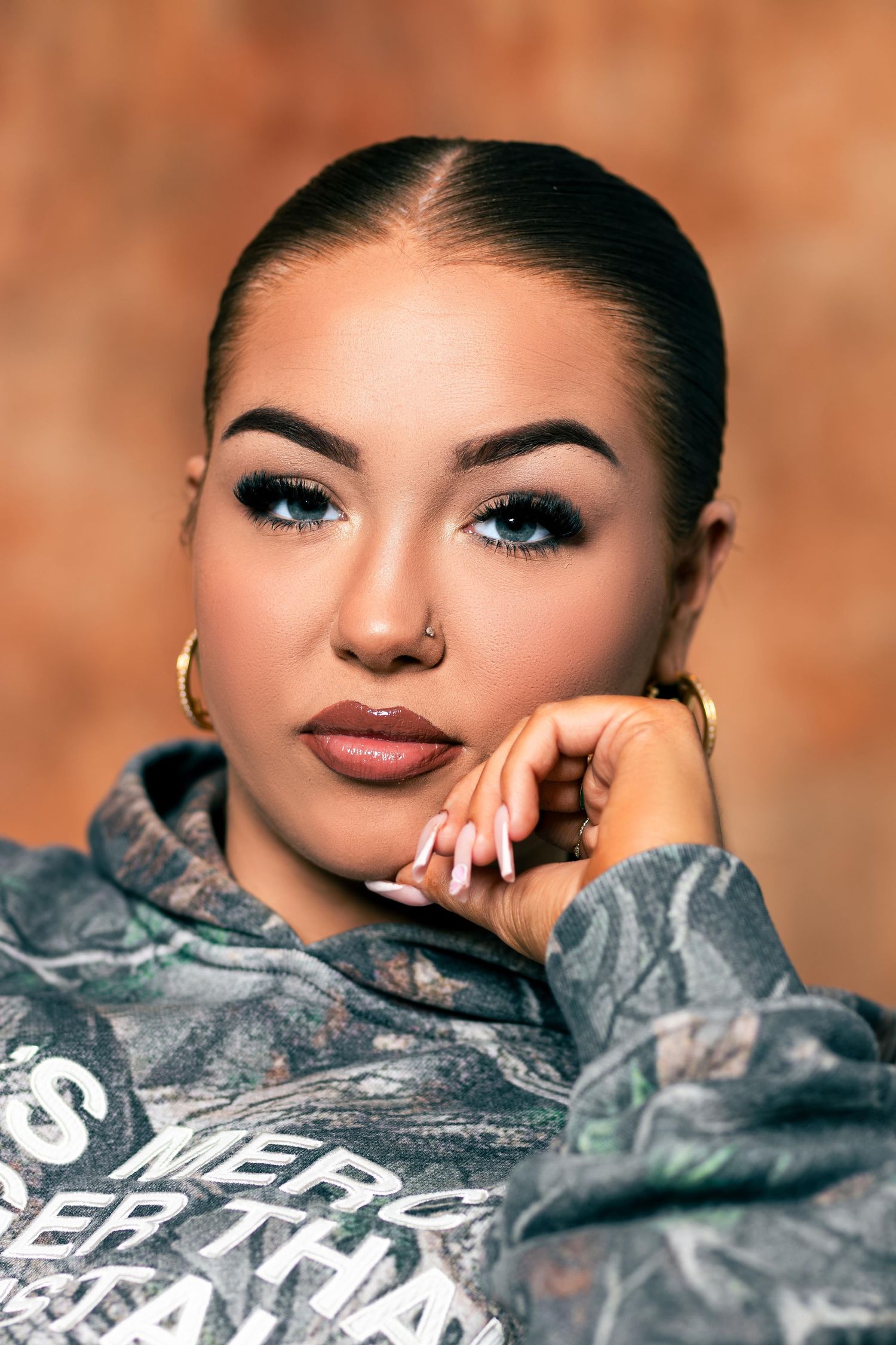 Young woman with sleek updo, bold eye makeup, nose ring, gold hoop earring, and camo hoodie posing against brown background.