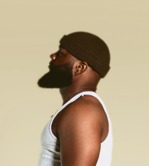 Side profile of bearded man in white tank top and olive beanie against neutral background.