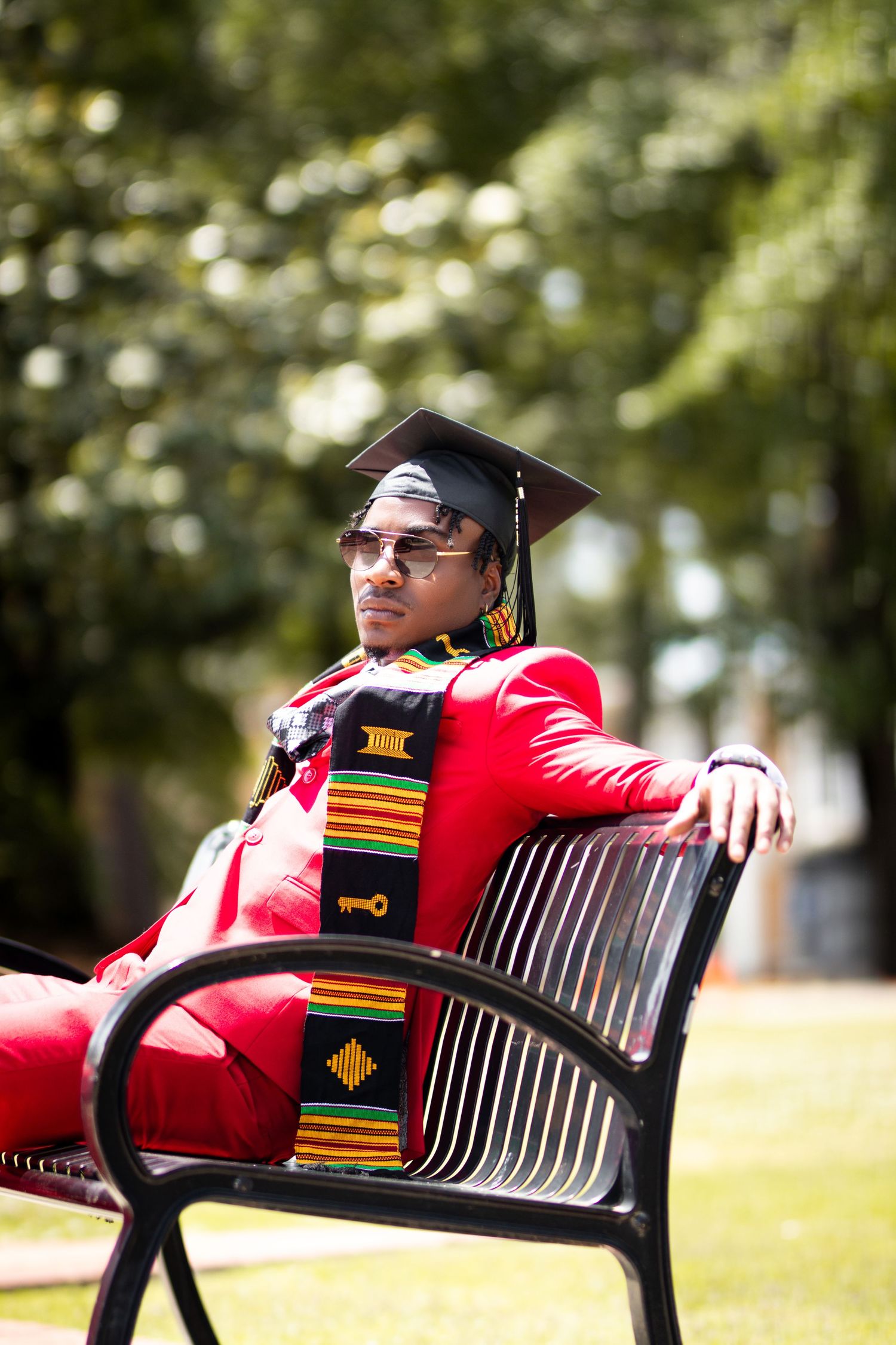 Young man in red suit and graduation cap with kente cloth stole seated on bench outdoors.