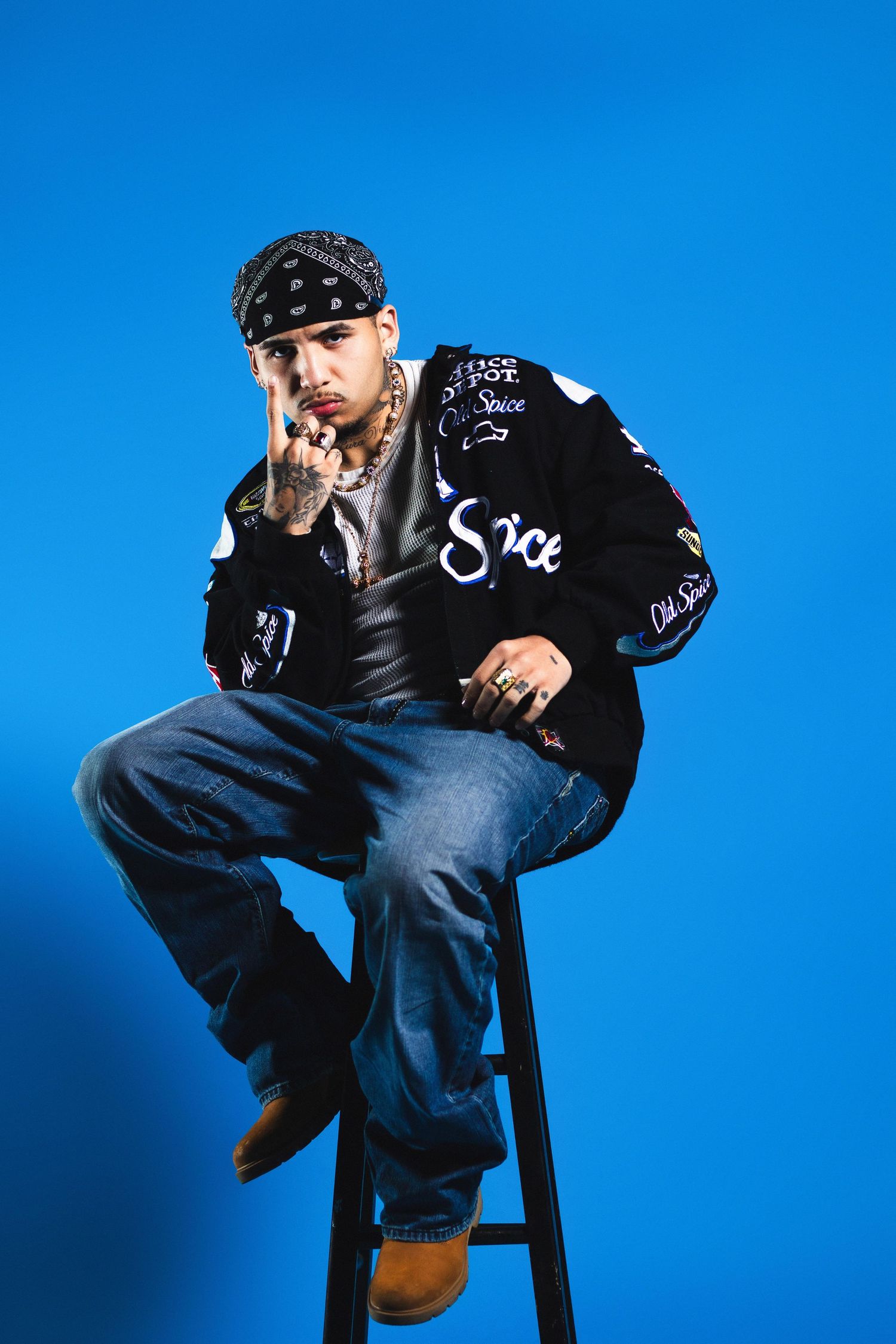 A tattooed Latino man in a bandana and branded jacket sits on a stool against a blue background.