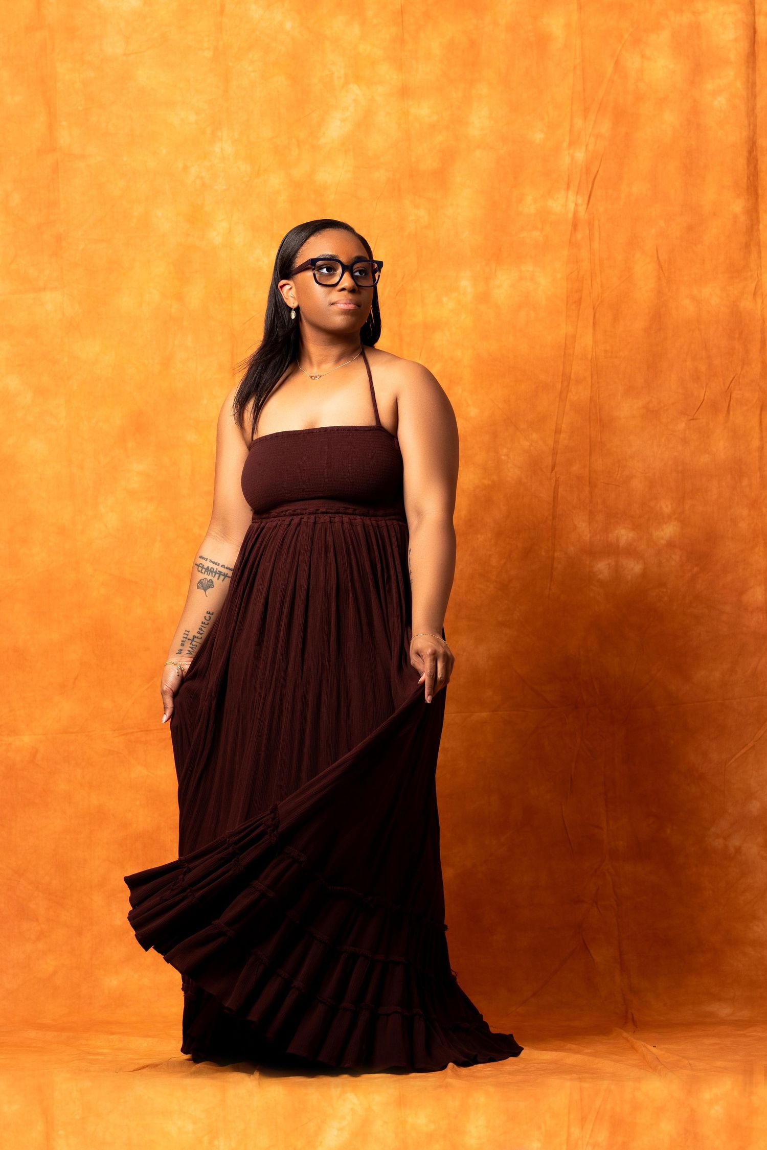 Woman in dark maroon maxi dress with glasses posing against vibrant orange textured background.