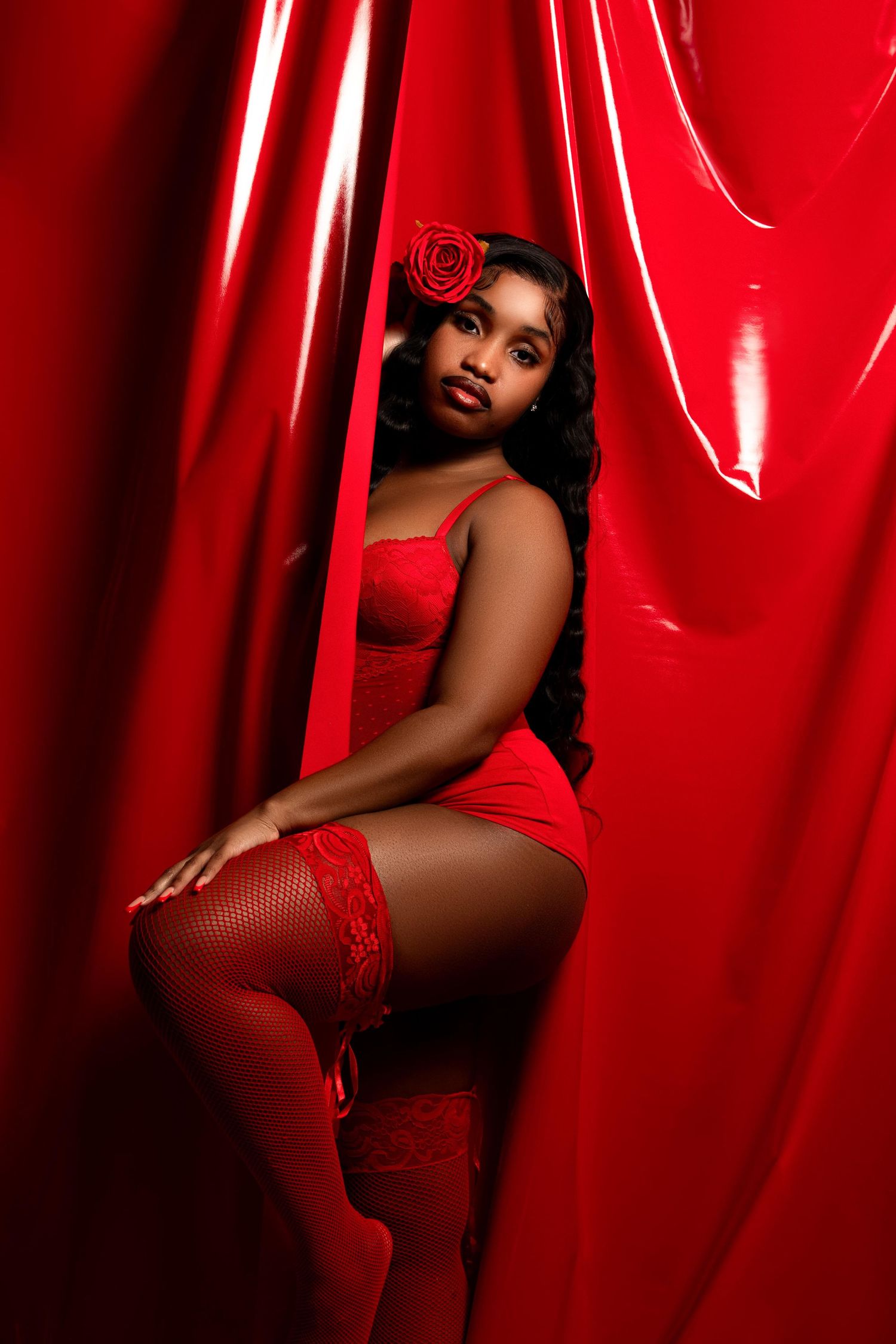 Woman in red lingerie and stockings posing by shiny red curtains with a rose hair accessory.