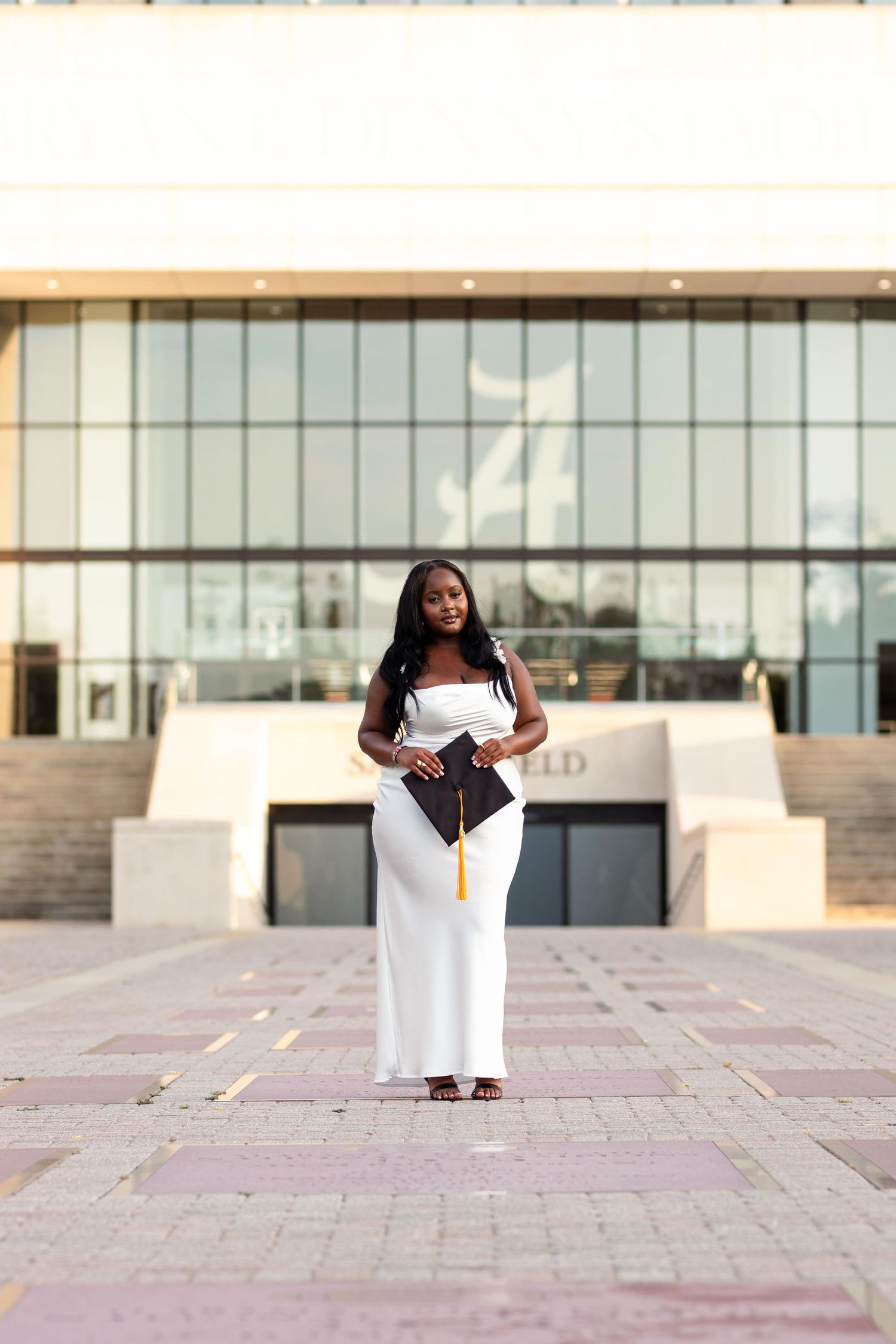 Young Black woman in white gown holding graduation cap outside modern university building.