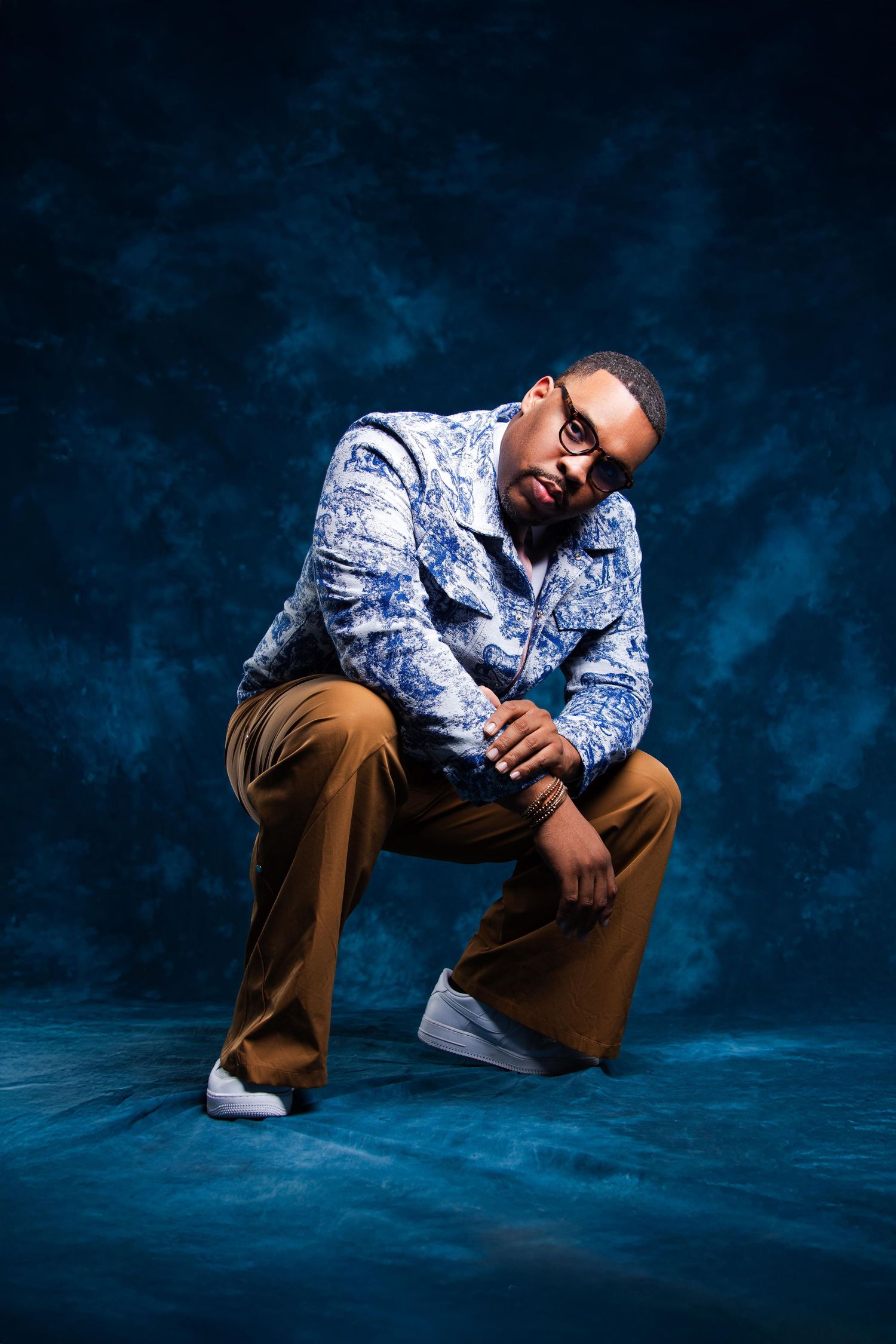 Man in blue floral shirt and tan pants kneeling confidently against a dark blue studio backdrop.