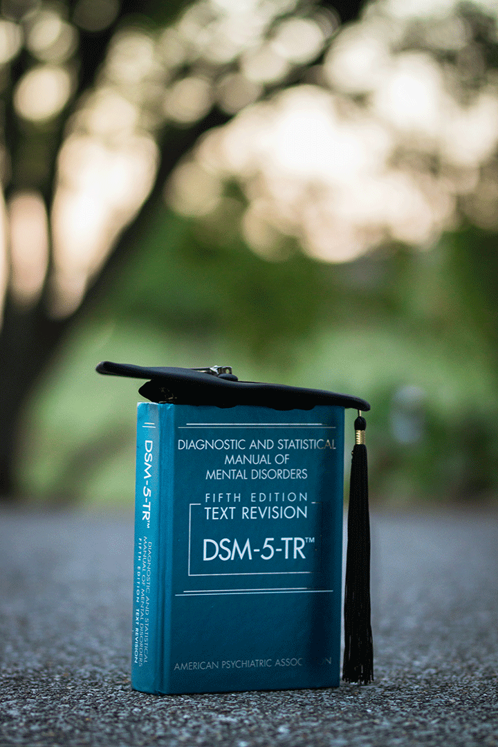 DSM-5-TR textbook topped with a graduation cap on pavement outdoors.