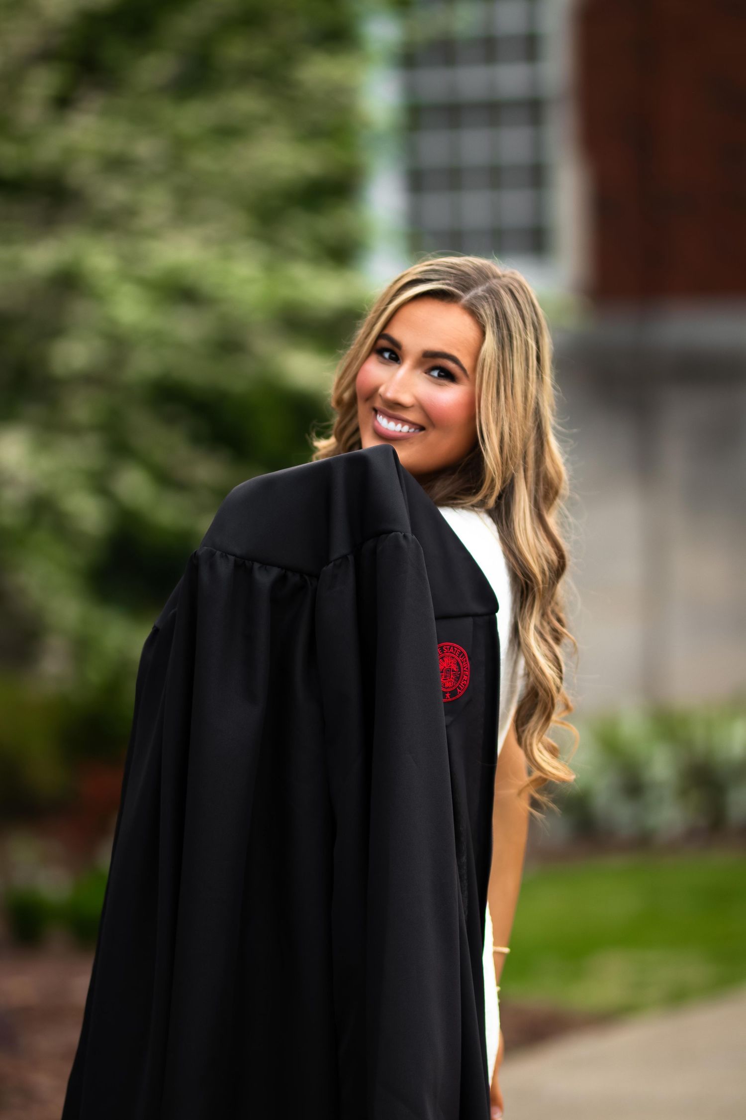 Smiling female graduate in black cap and gown posing outdoors on campus with lush green trees.