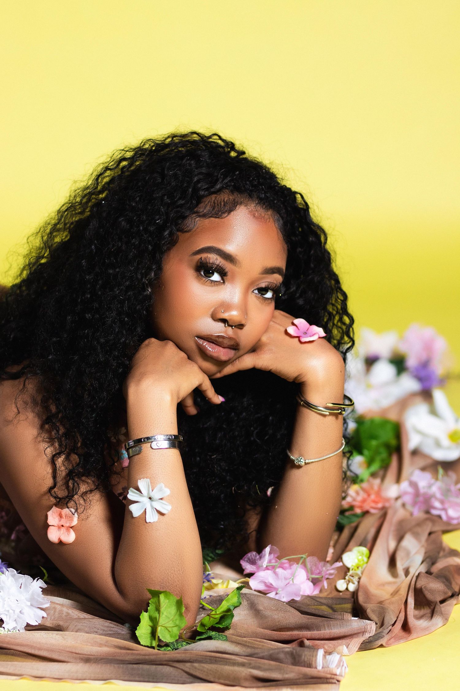 A young Black woman with curly hair lies among flowers wearing bracelets against a yellow background.