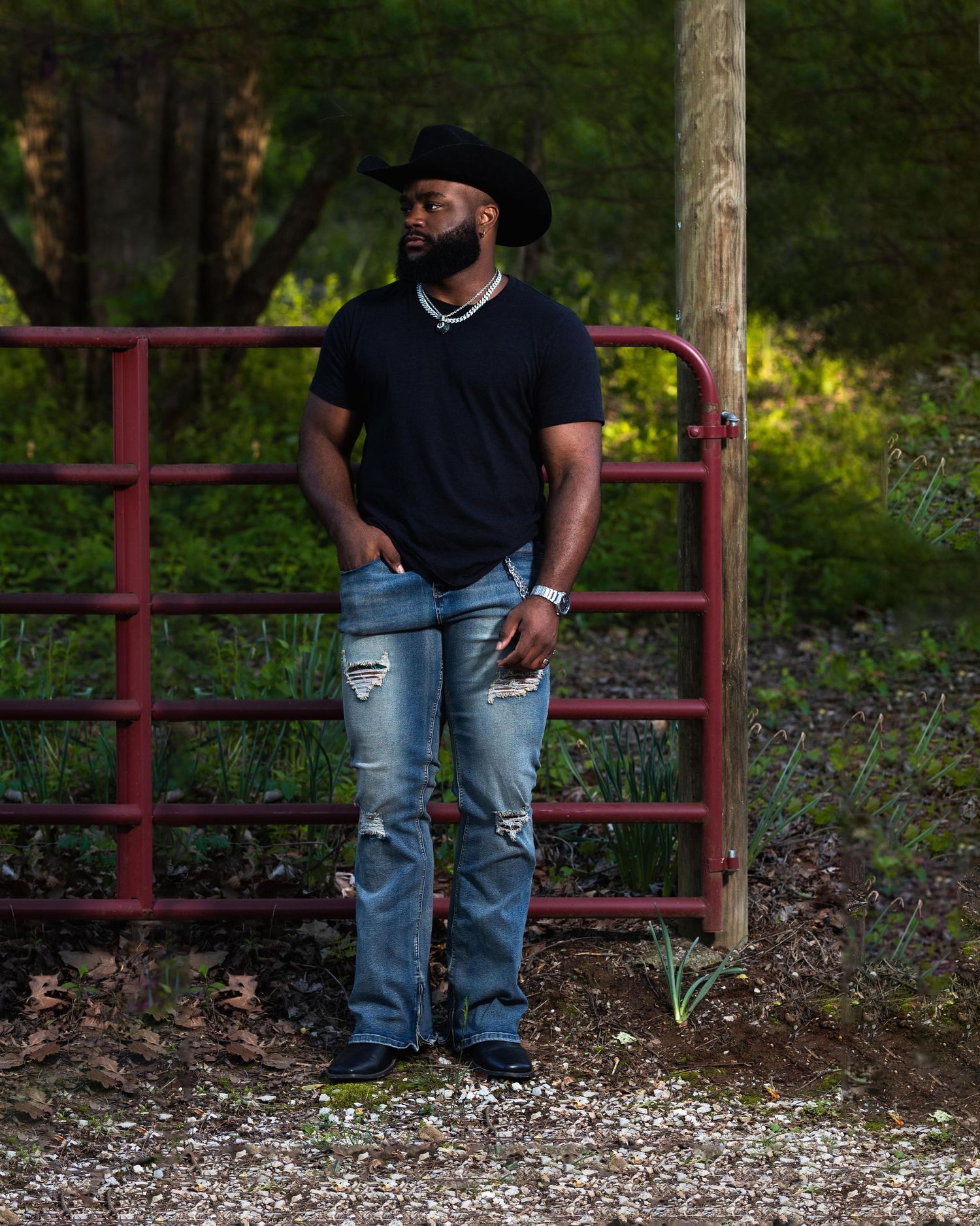 Man in black cowboy hat and ripped jeans leaning on red farm gate outdoors.