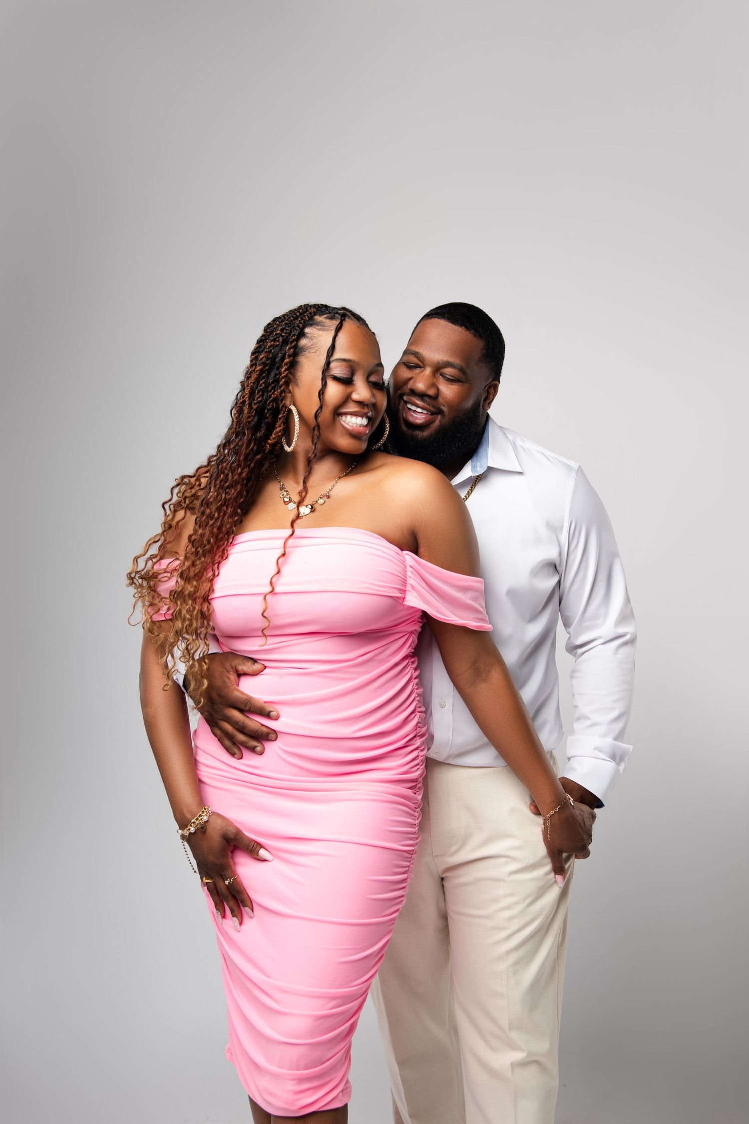 A joyful Black couple poses together, woman in pink off-shoulder dress, man in white shirt and cream pants, gray background.
