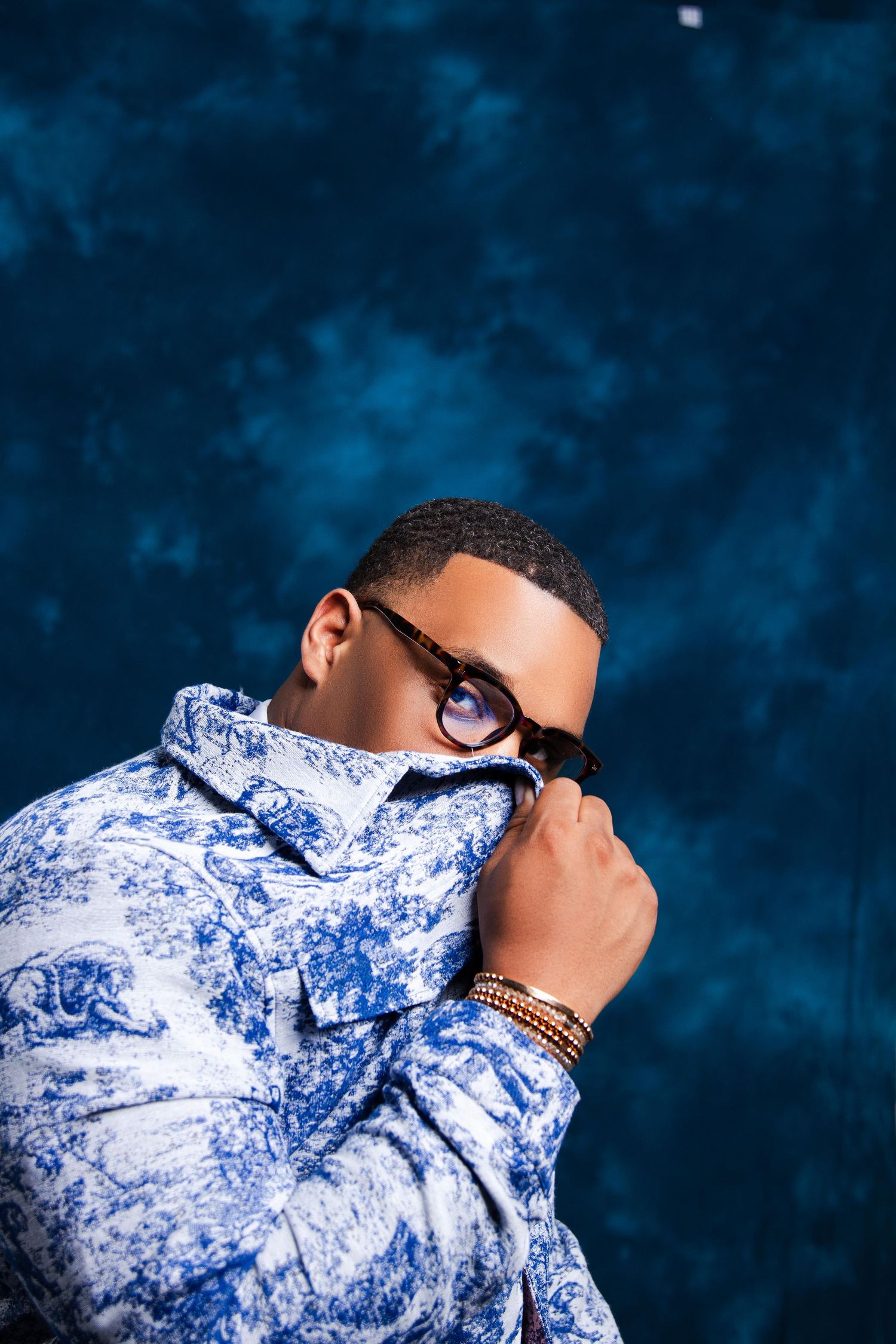 Man in blue floral shirt and glasses posing against dark blue background, wearing beaded bracelets.
