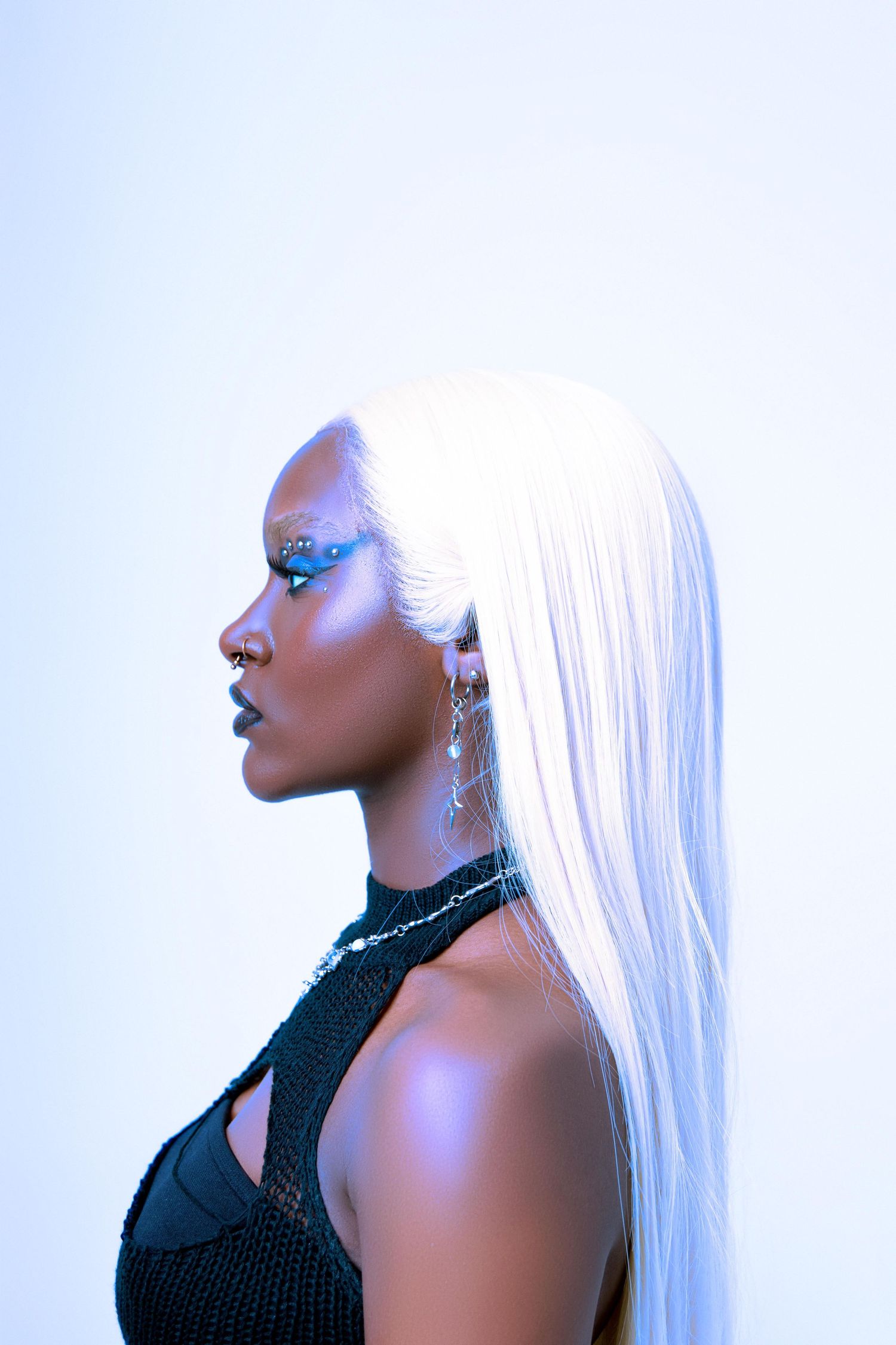 A Black woman with a long white wig and blue eye makeup poses in profile against a light background.