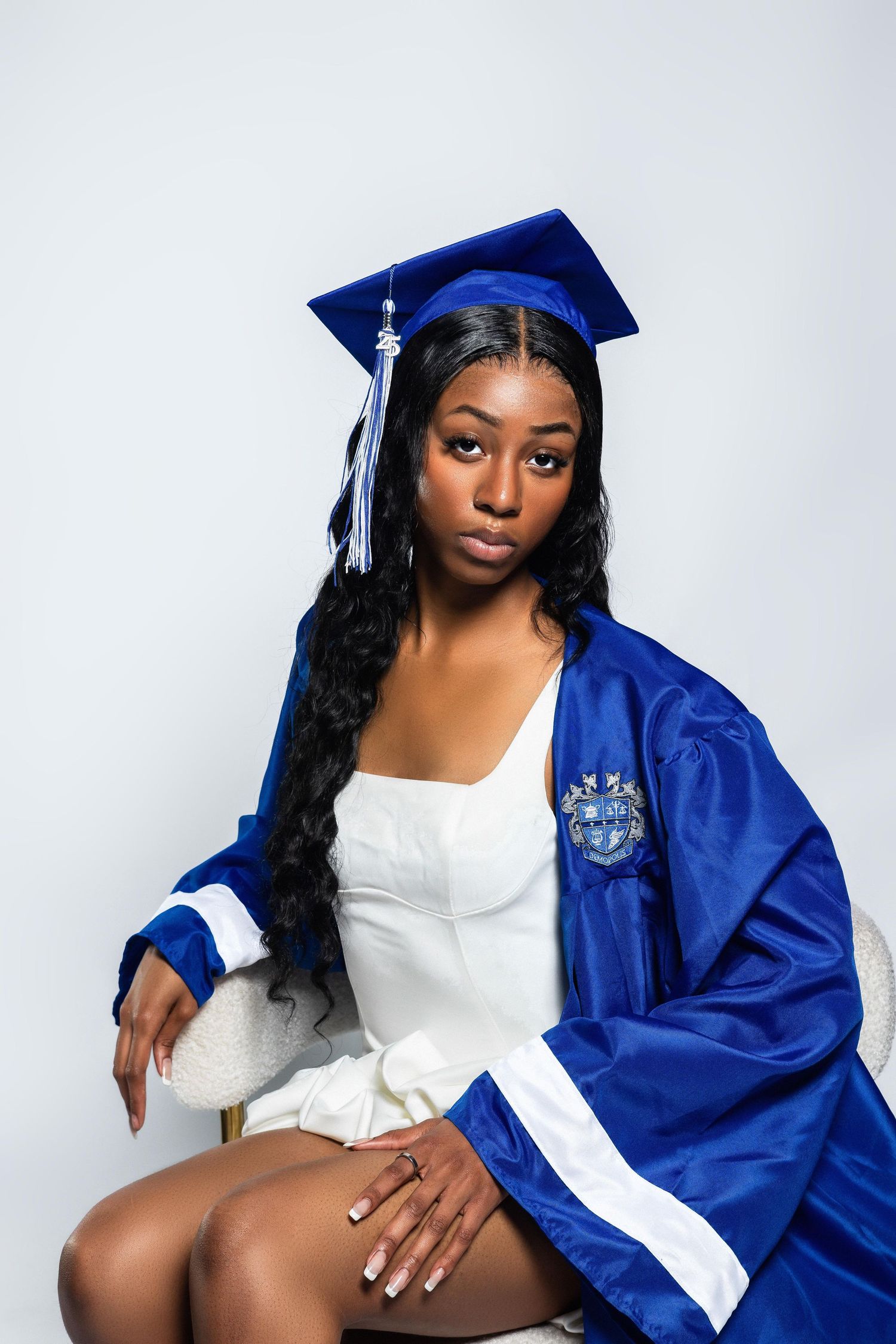 Young Black woman in blue graduation cap and gown seated confidently on a chair in studio.