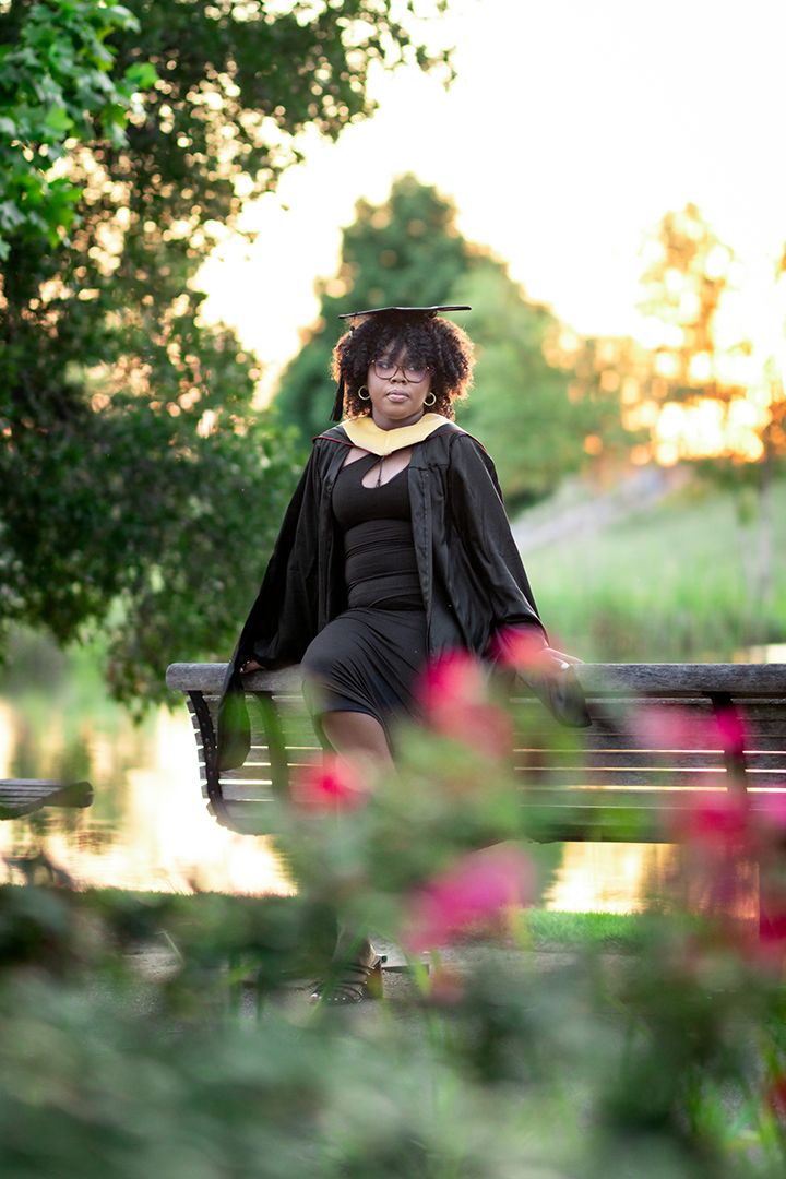 Female graduate in black cap and gown sits on bench near pond at sunset.
