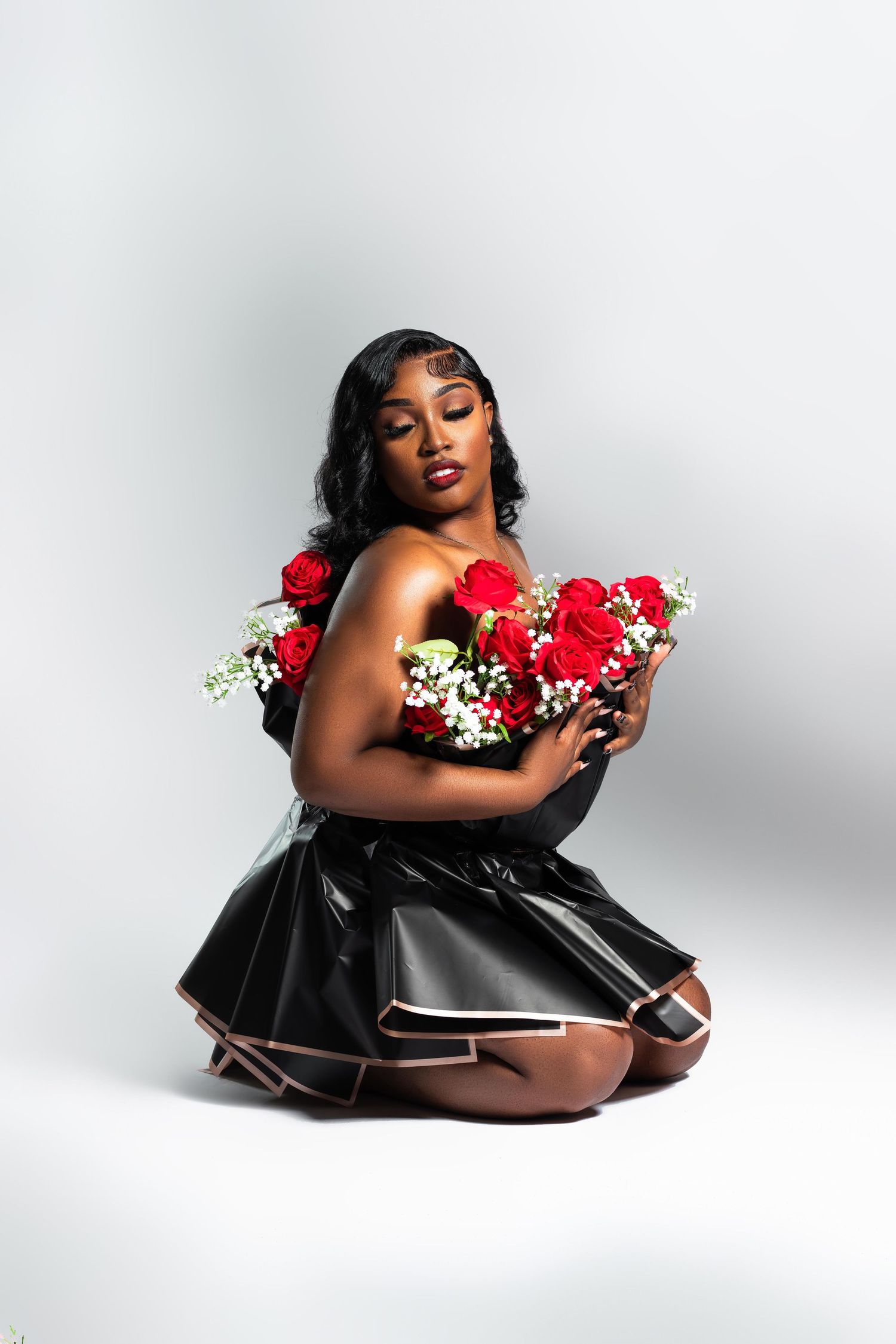 A Black woman in black leather dress kneels holding red roses and white flowers on gray background.